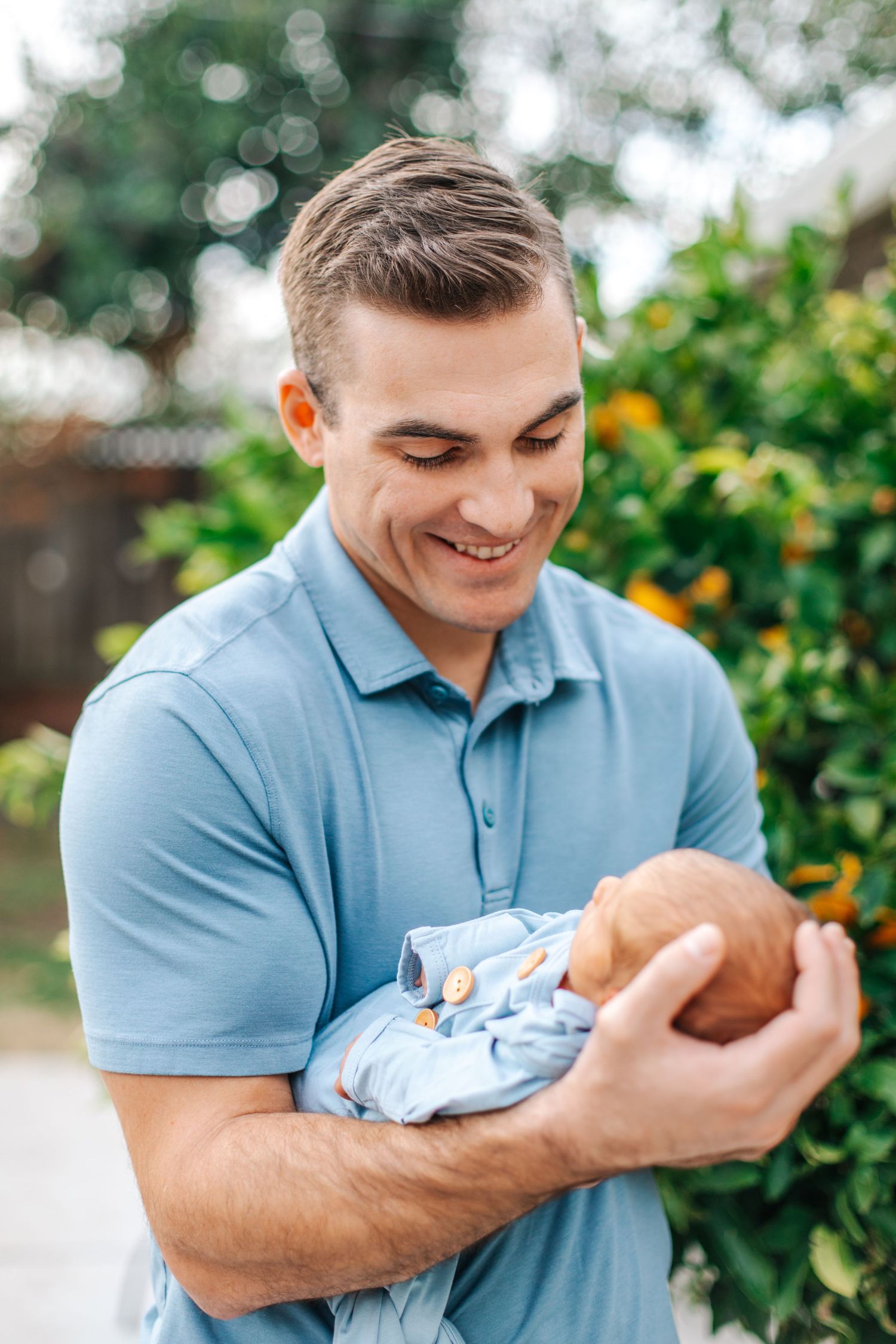 Smiling father in blue polo shirt holding newborn baby outdoors near orange tree.