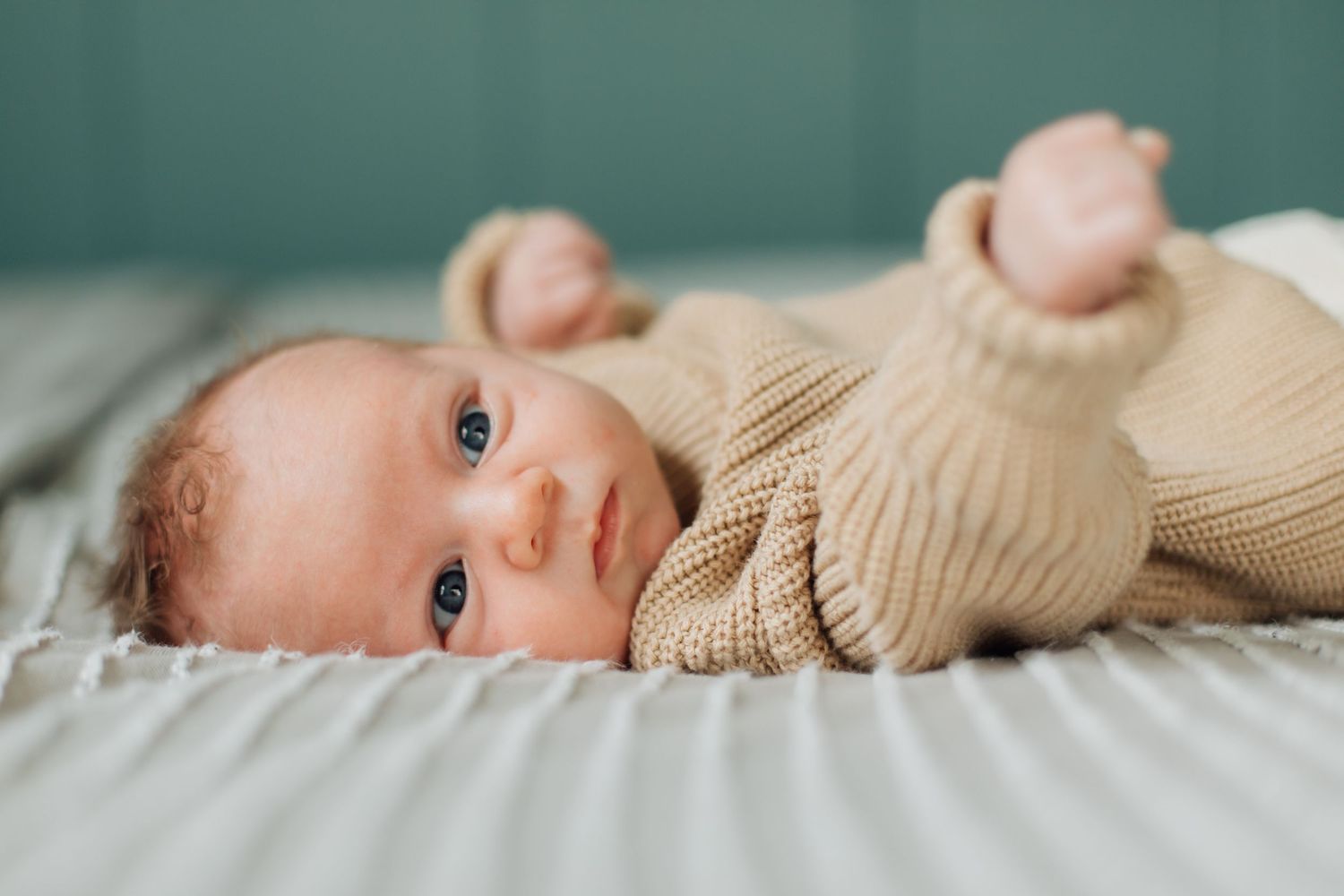 A newborn baby in a beige knit outfit lies on a bed, gazing upward with bright blue eyes.
