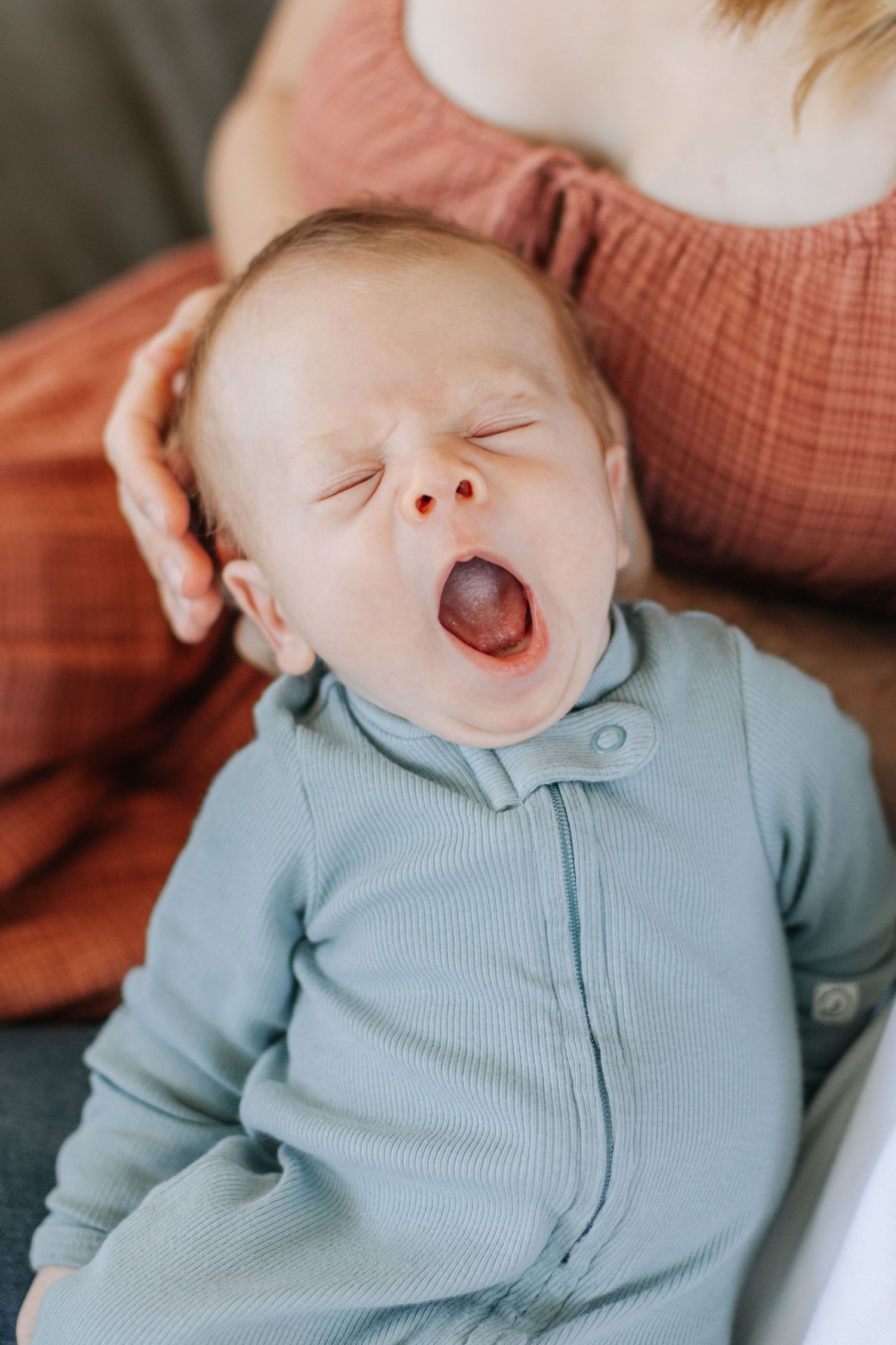 Close-up of a newborn baby yawning widely while wearing a light blue ribbed zip-up onesie.