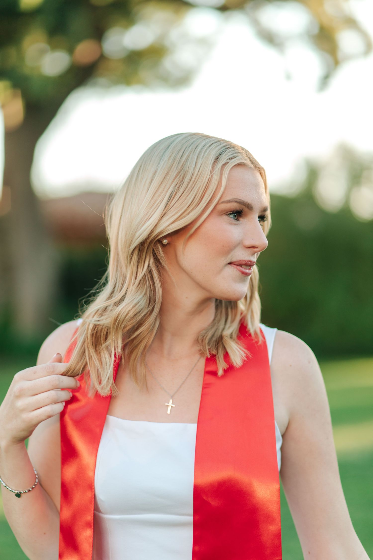 Close-up of blonde woman wearing red graduation stole and cross necklace outdoors in sunlight.