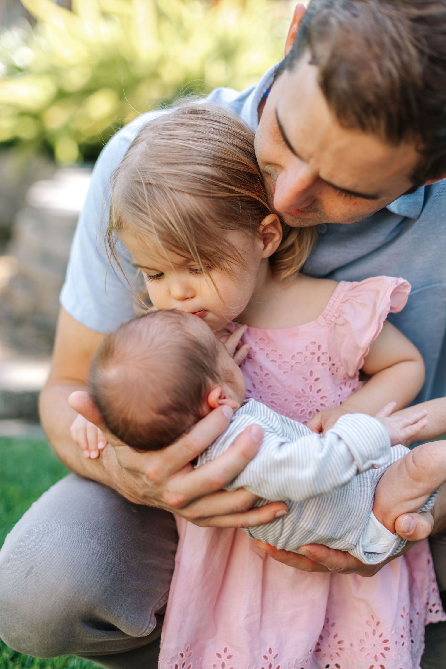 Father crouching outdoors while toddler girl in pink dress kisses newborn sibling he is holding.