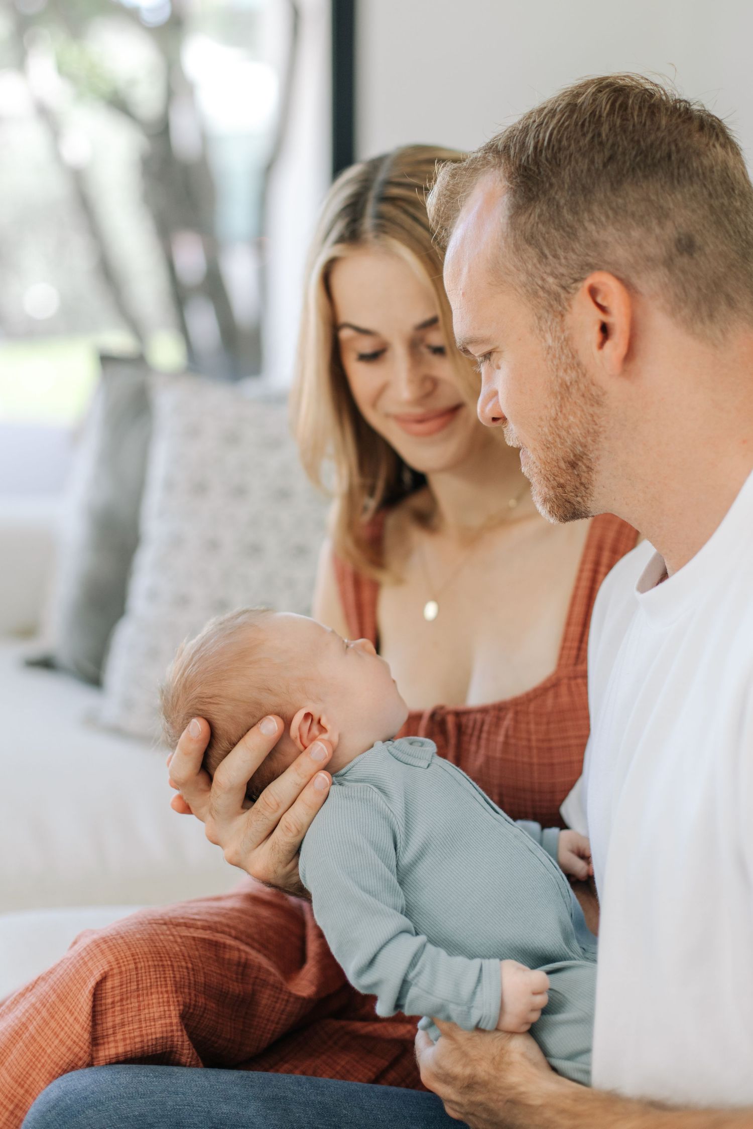 New parents gazing lovingly at their newborn baby dressed in a light blue onesie on a sofa.