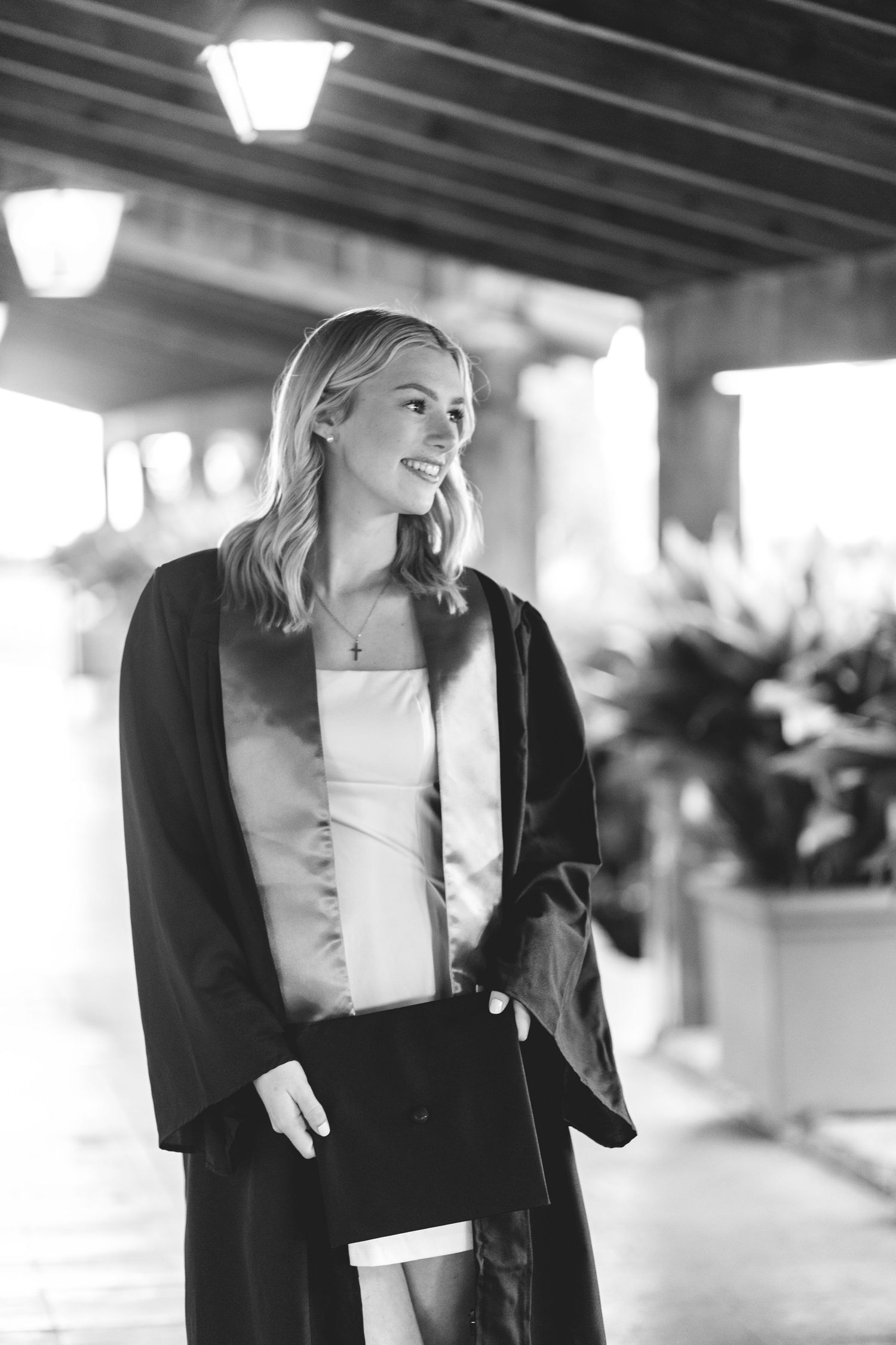 Black and white photo of smiling blonde woman in graduation gown and stole under lit walkway.