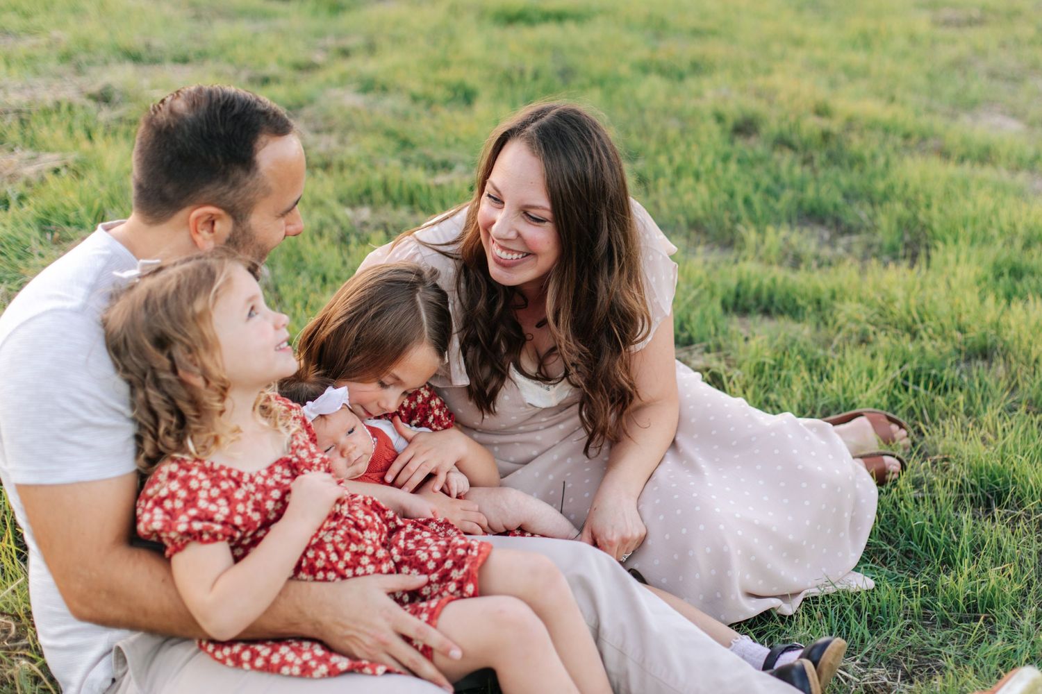 A happy family of four sits together on green grass, laughing and enjoying time outdoors.