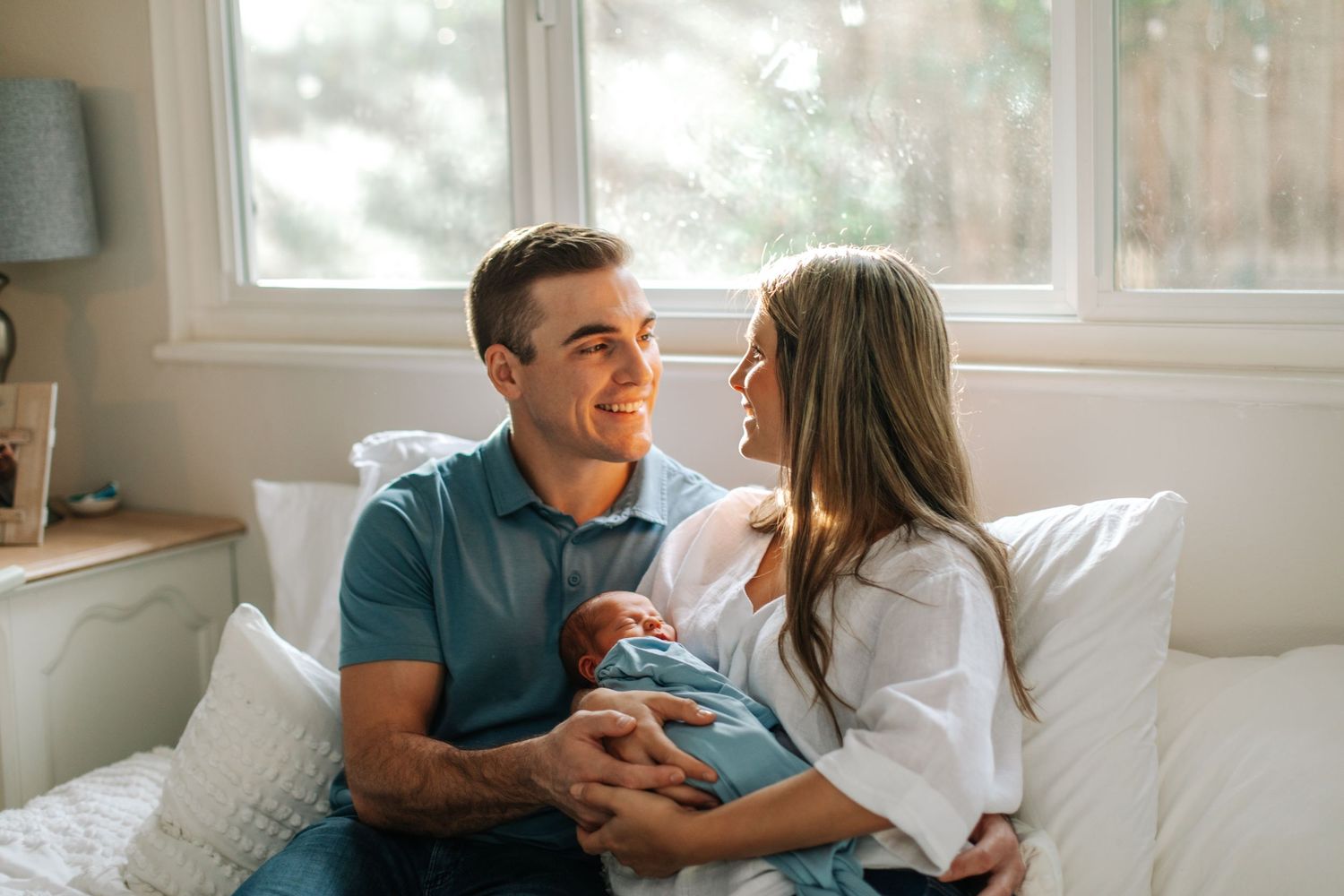 Happy couple sitting on bed smiling at each other while holding their newborn baby.