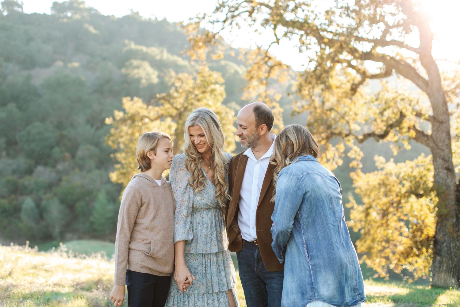 Family of four laughing together outdoors in golden sunlight beneath oak trees.