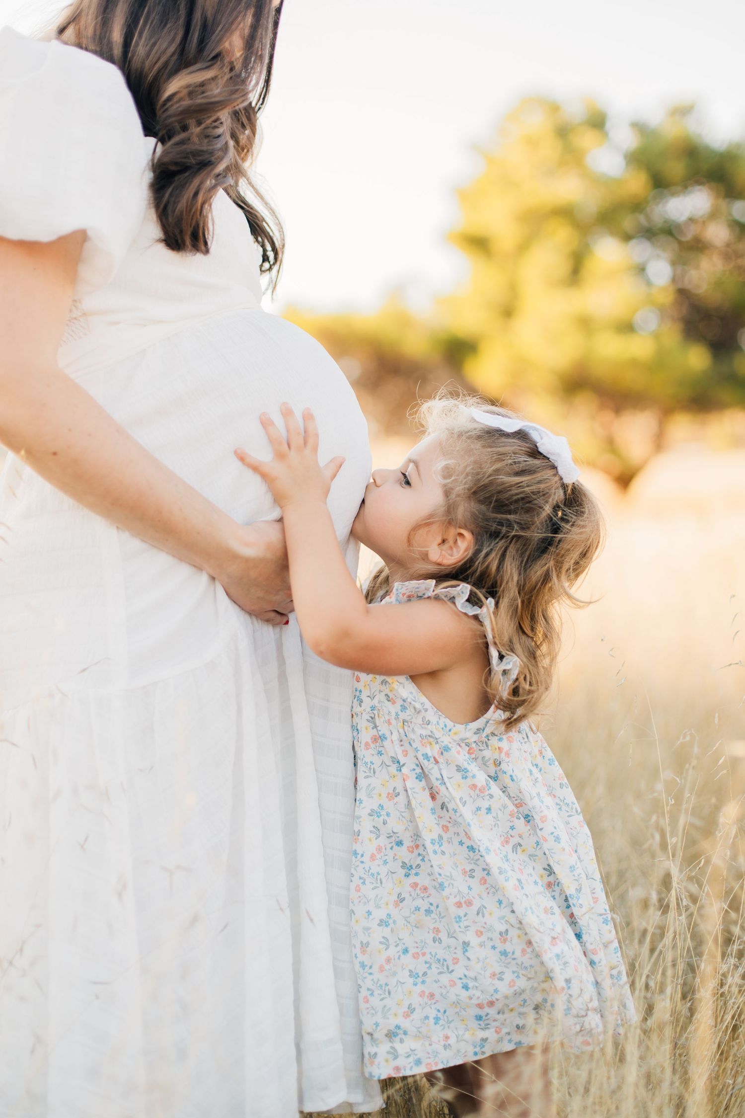 A young girl in a floral dress kisses her pregnant mother's belly in a sunny field.