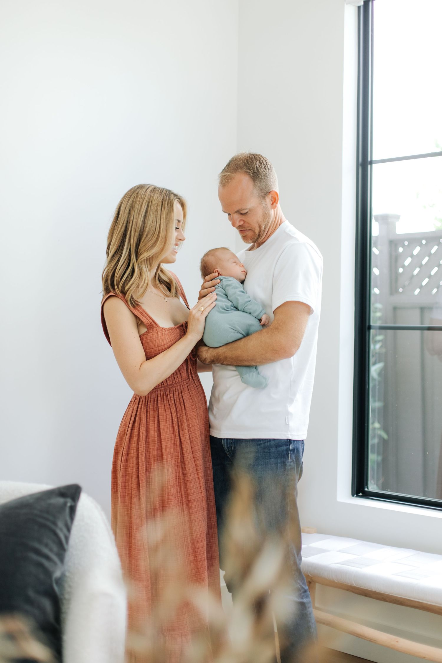 Young couple in rust and white outfits standing by window admiring newborn baby together.