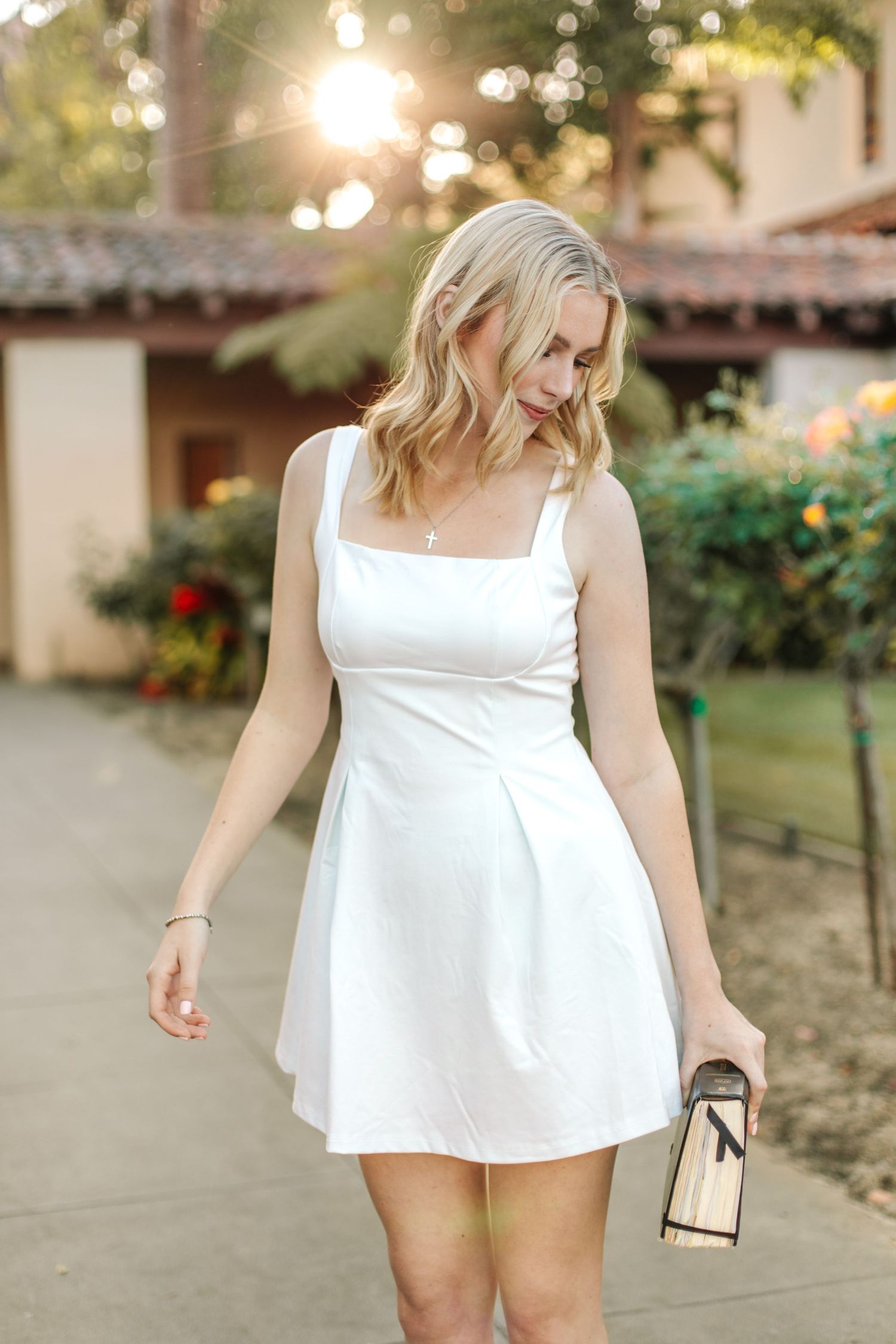 Blonde woman in white sundress walks through garden holding graduation diploma in golden light.