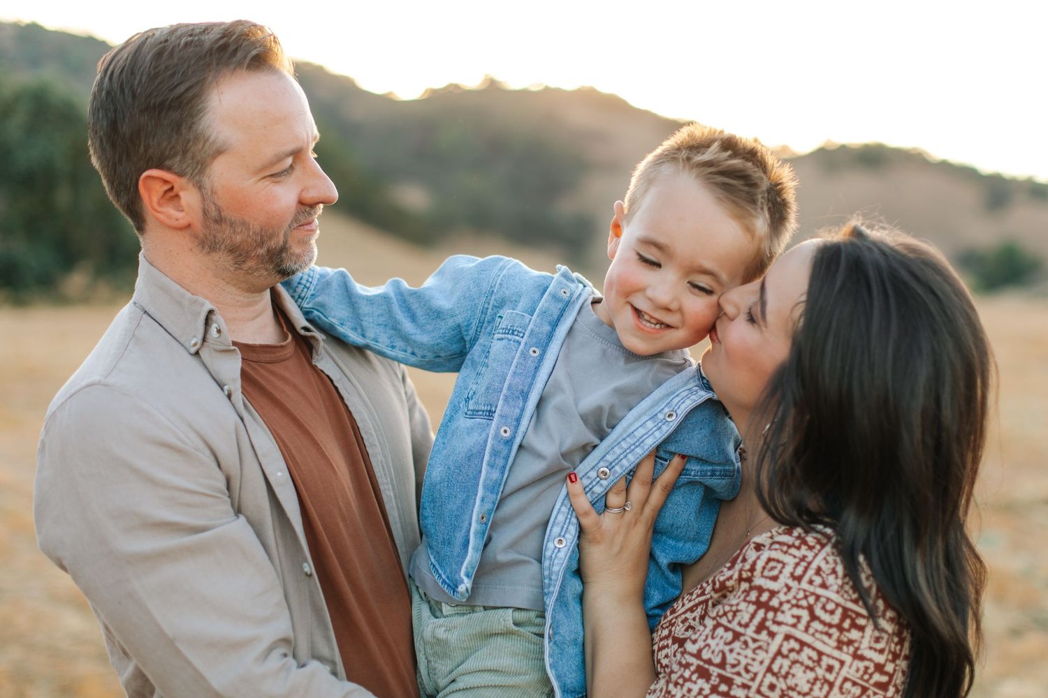Parents lovingly kissing their happy toddler son outdoors in a sunny golden field.