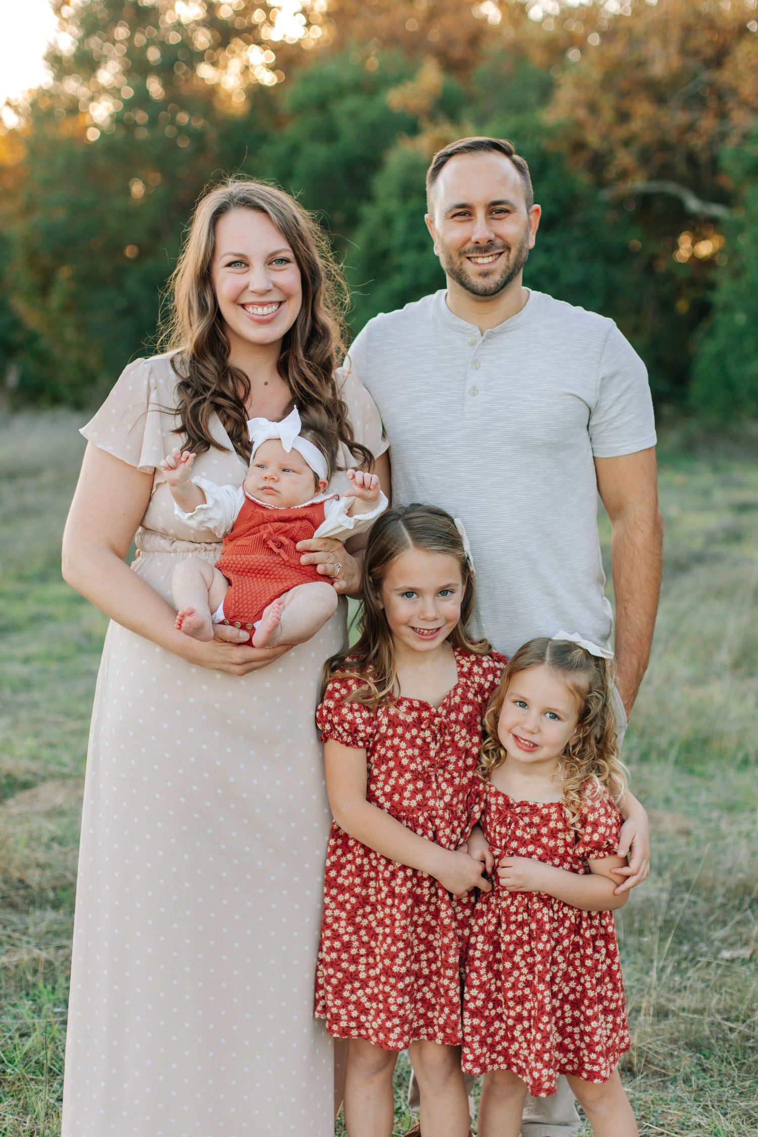 A smiling family of five poses outdoors at golden hour, with three daughters in matching red dresses.