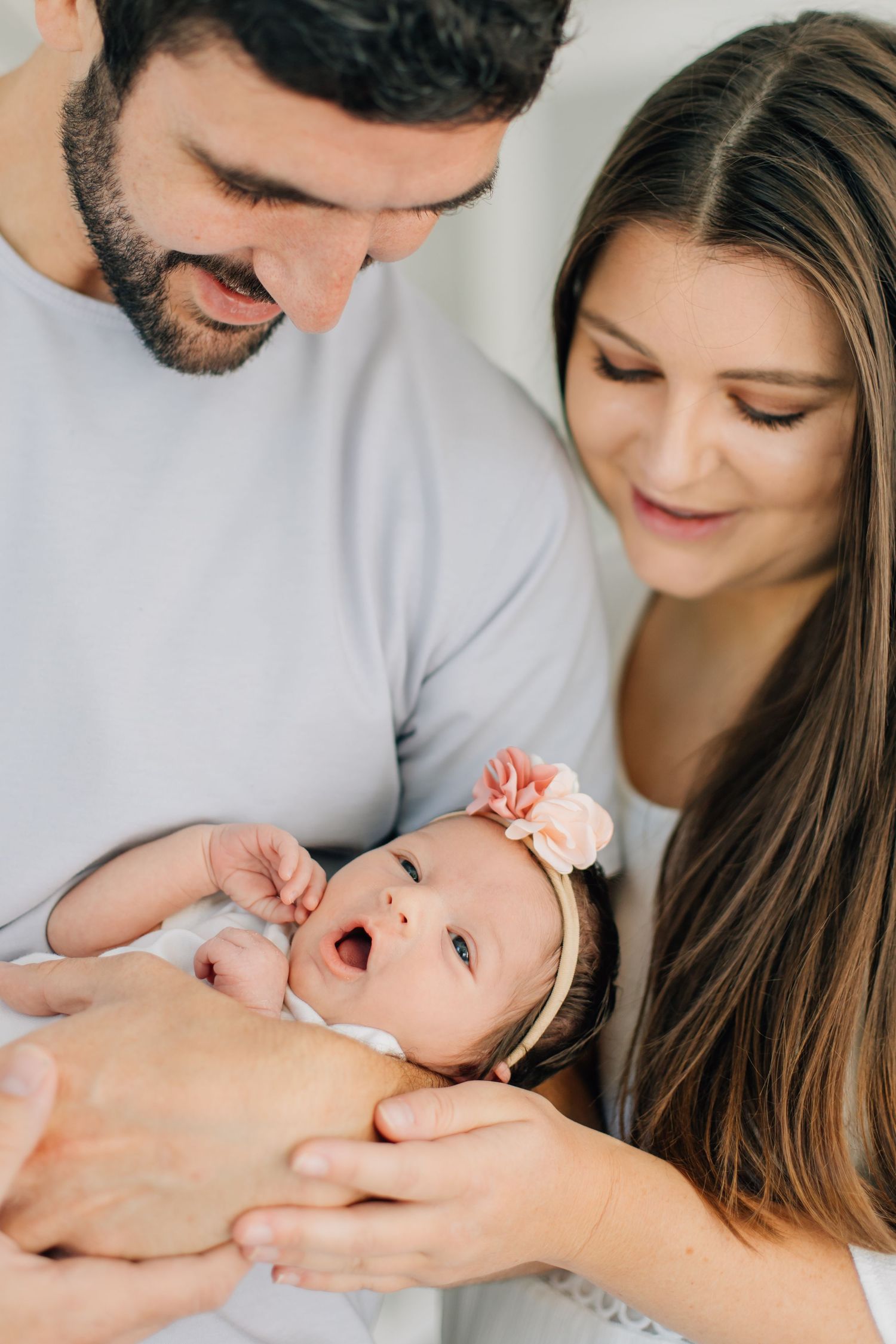 Young parents lovingly look at their wide-eyed newborn baby girl with pink flower headband.
