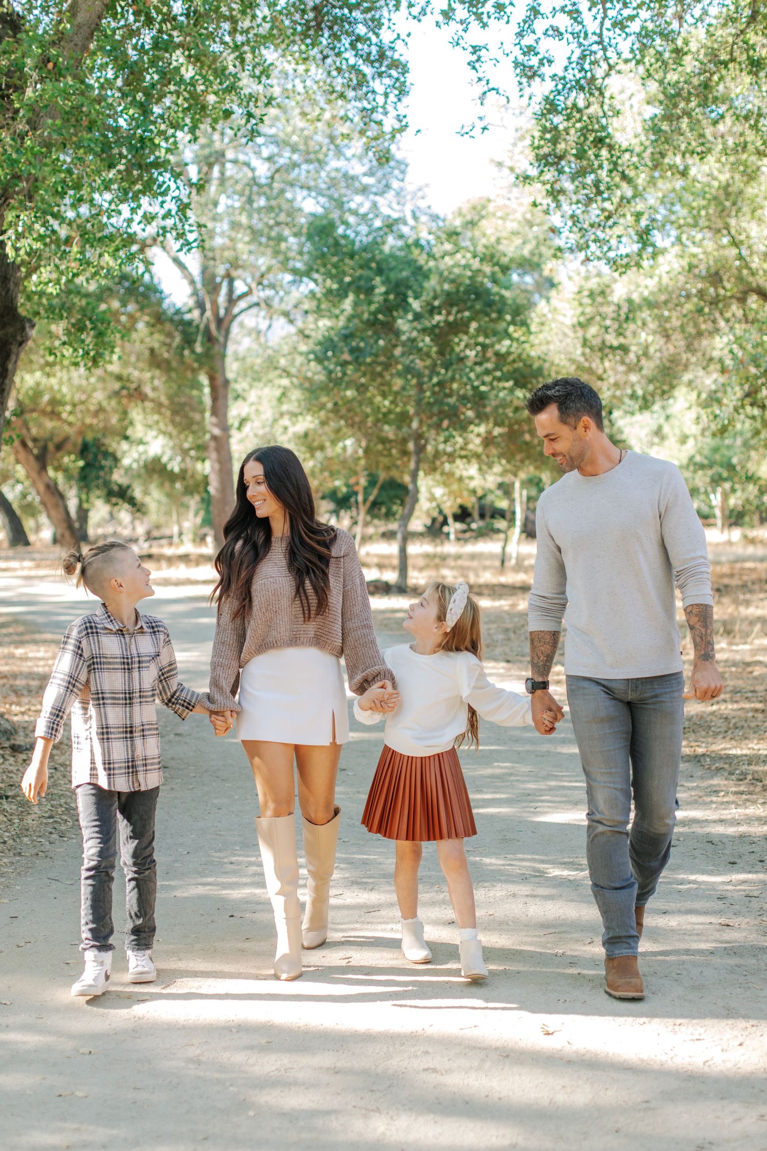 Happy family of four walking hand in hand along a sunny tree-lined path.
