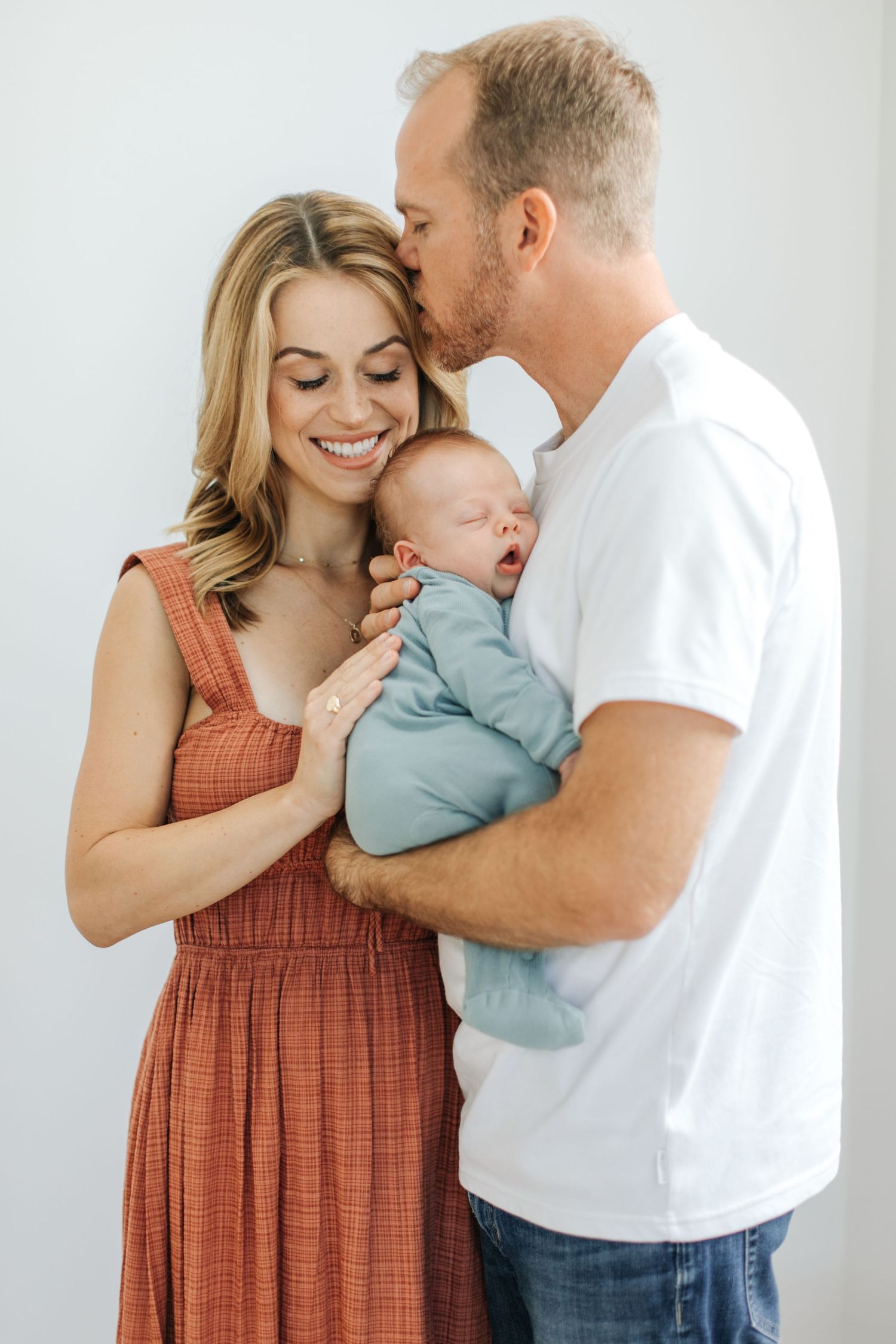 Smiling couple in rust and white outfits holding yawning newborn baby in blue onesie.