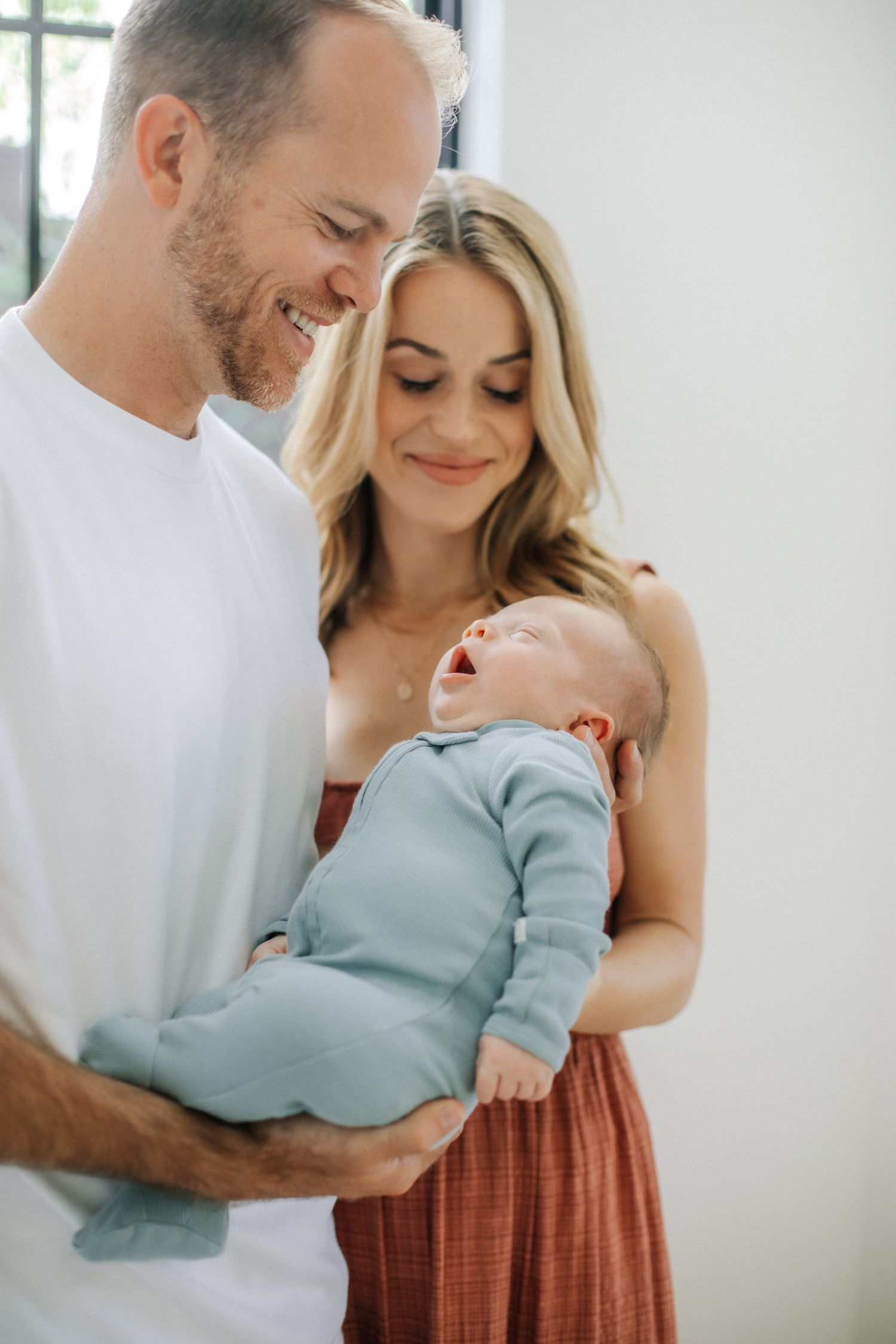 Smiling parents holding yawning newborn baby in light blue outfit in bright white room.