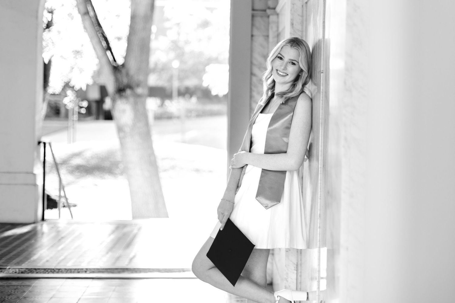 Black and white photo of blonde graduate in white dress leaning against column holding cap.