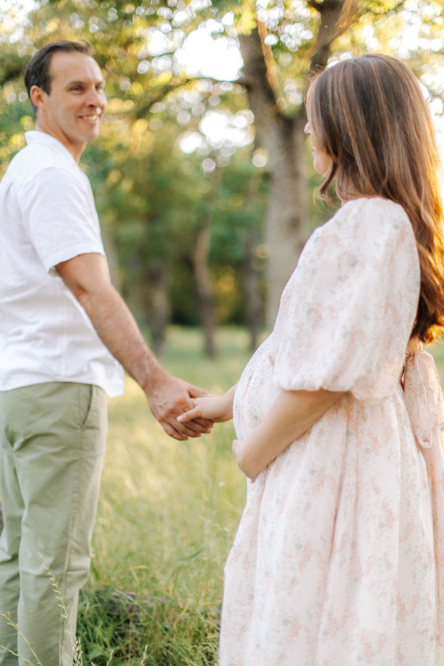 Smiling man holding hands with pregnant woman in floral dress in sunlit green meadow.