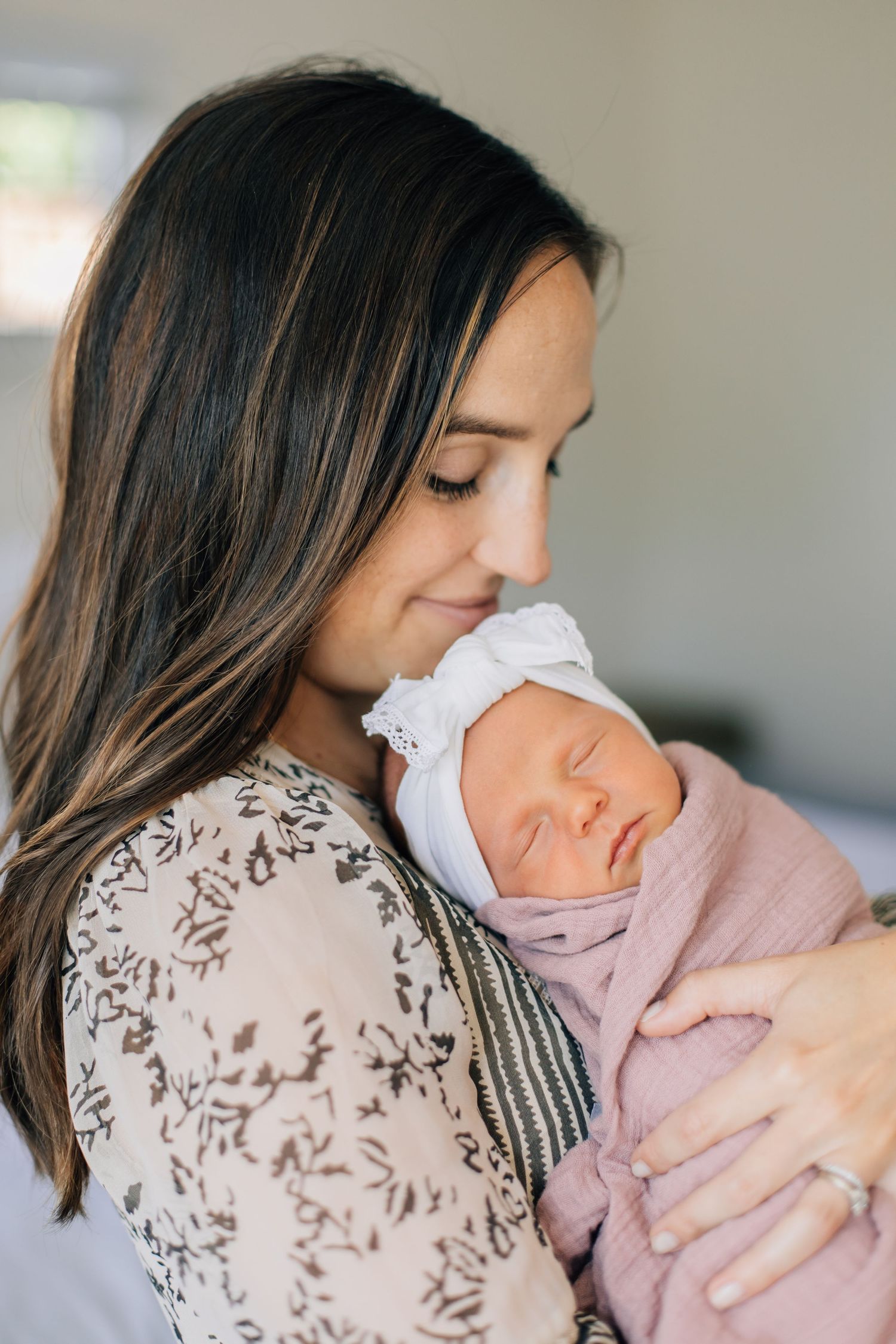 Mother with long dark hair holds sleeping newborn wrapped in pink swaddle with white lace headband.
