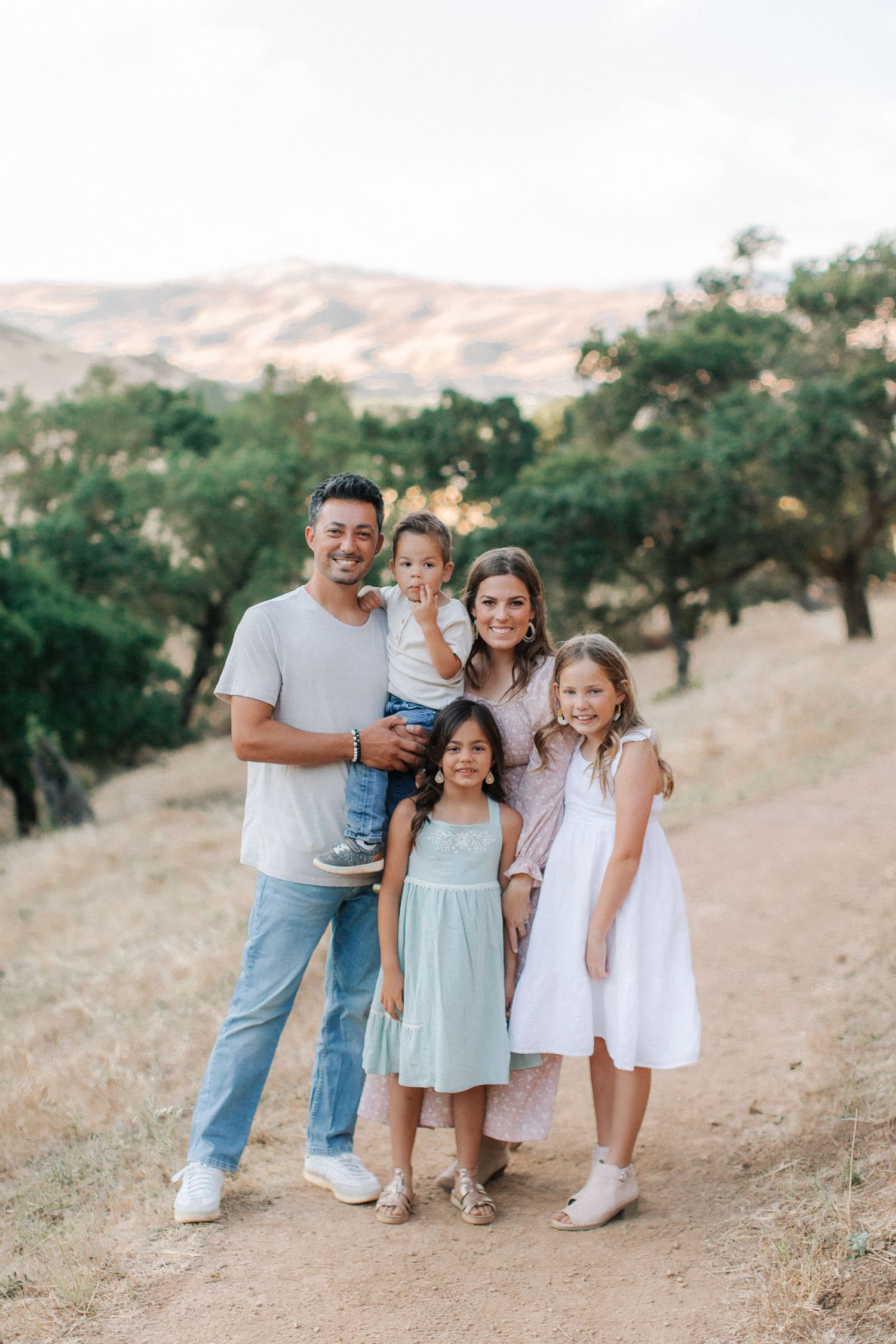 Happy family of five smiling together on a dirt path with trees and hills in background.