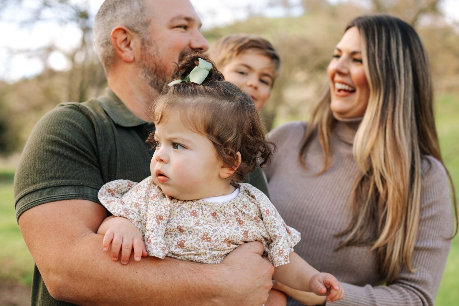 Happy family of four with a baby girl smiling together outdoors in a green park setting.