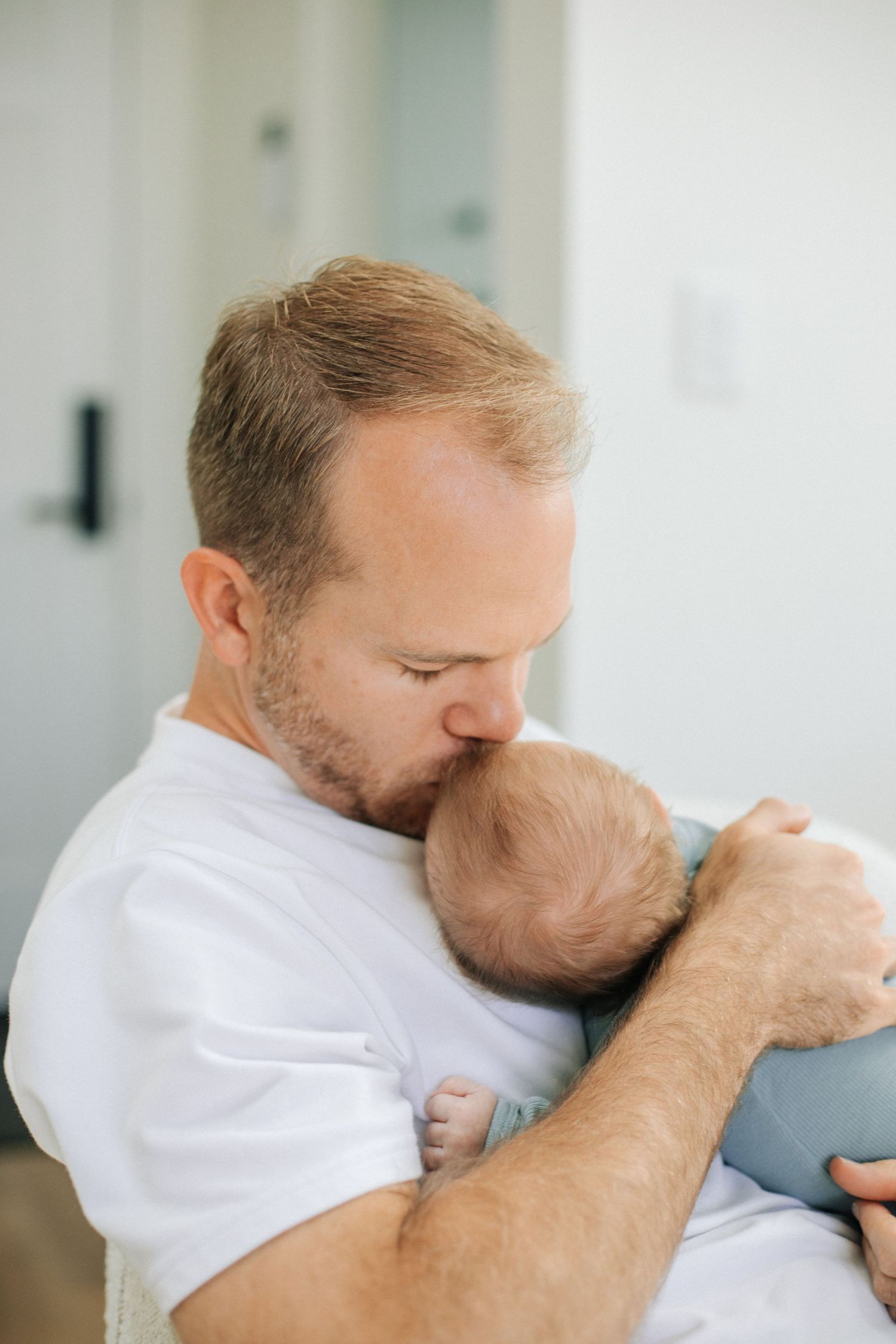 Father kissing newborn baby on head while holding infant gently in white t-shirt indoors.