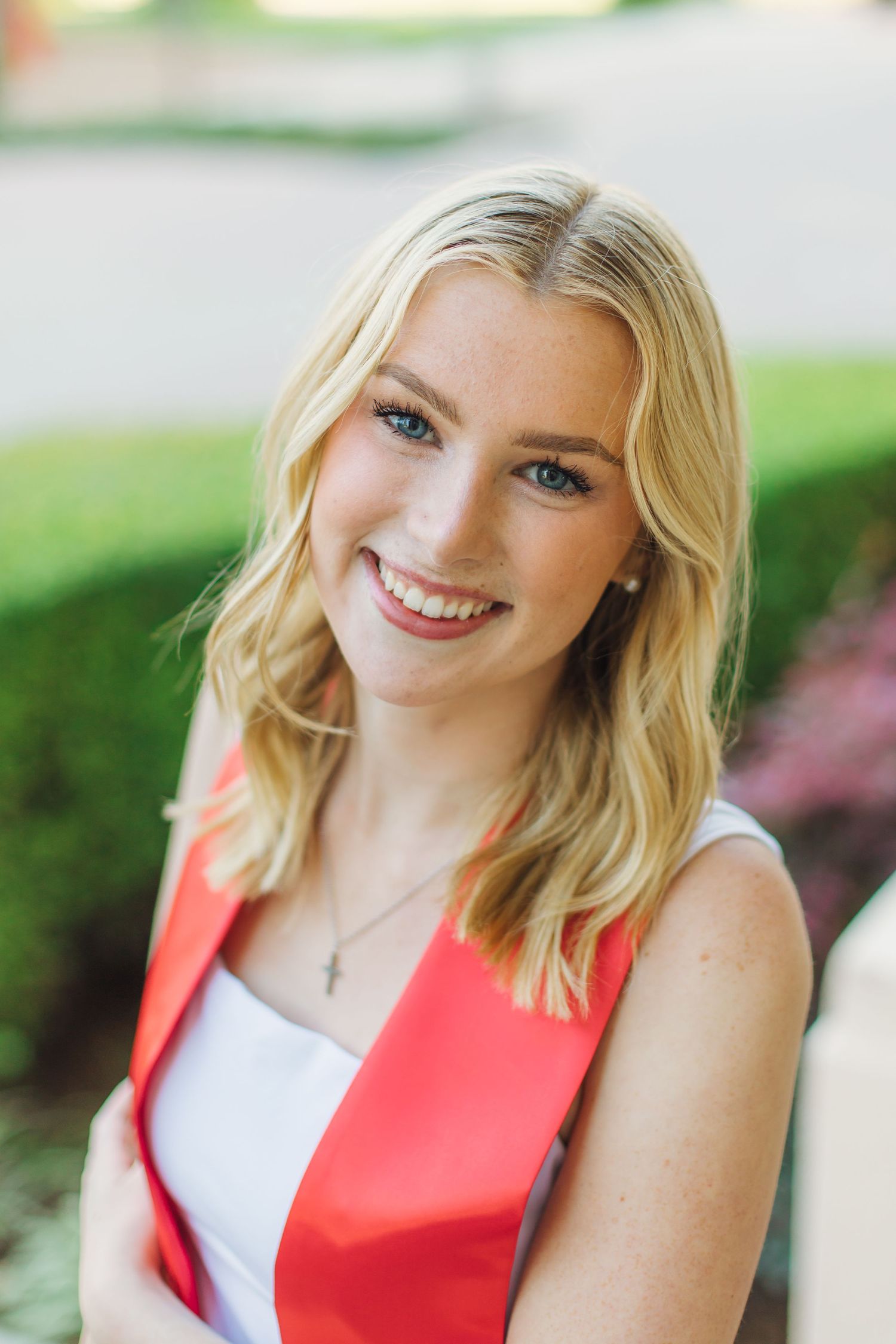 Close-up portrait of smiling blonde graduate wearing red stole and cross necklace outdoors.
