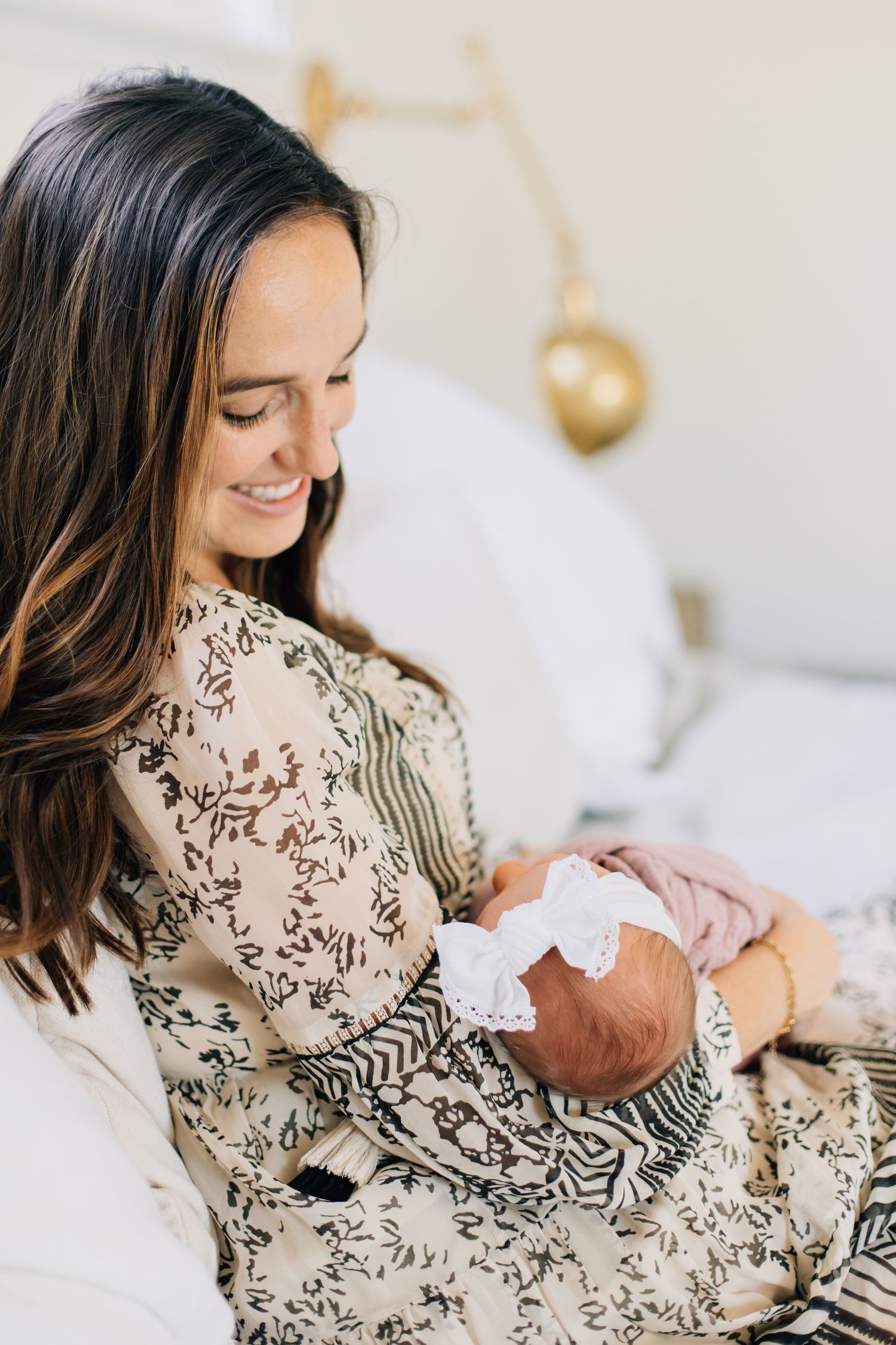 Smiling brunette mother in floral dress gazes lovingly at her newborn baby with white bow headband.