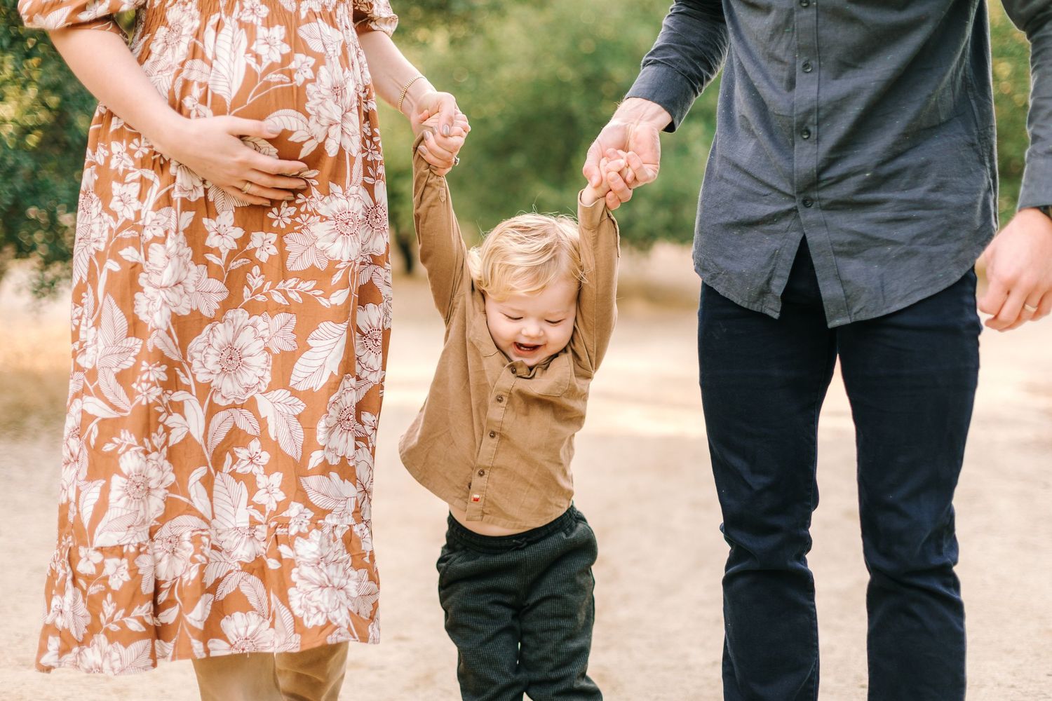 Laughing toddler held by hands between pregnant mother and father on a sunny outdoor path.