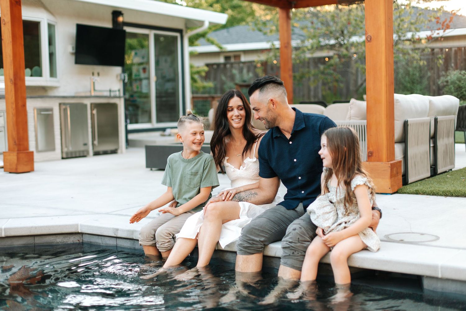 Family of four sitting poolside on a sunny patio, laughing and dipping feet in water.