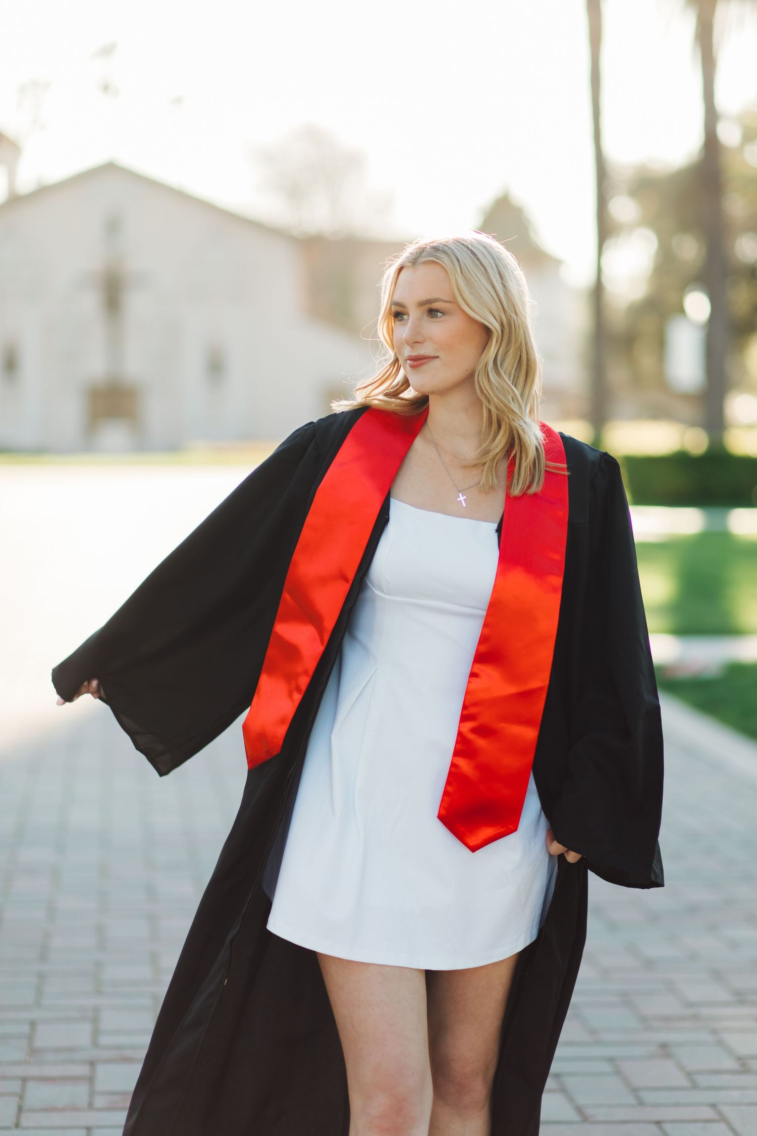 Blonde woman in black graduation gown with red stole walking on brick path near white mission church.