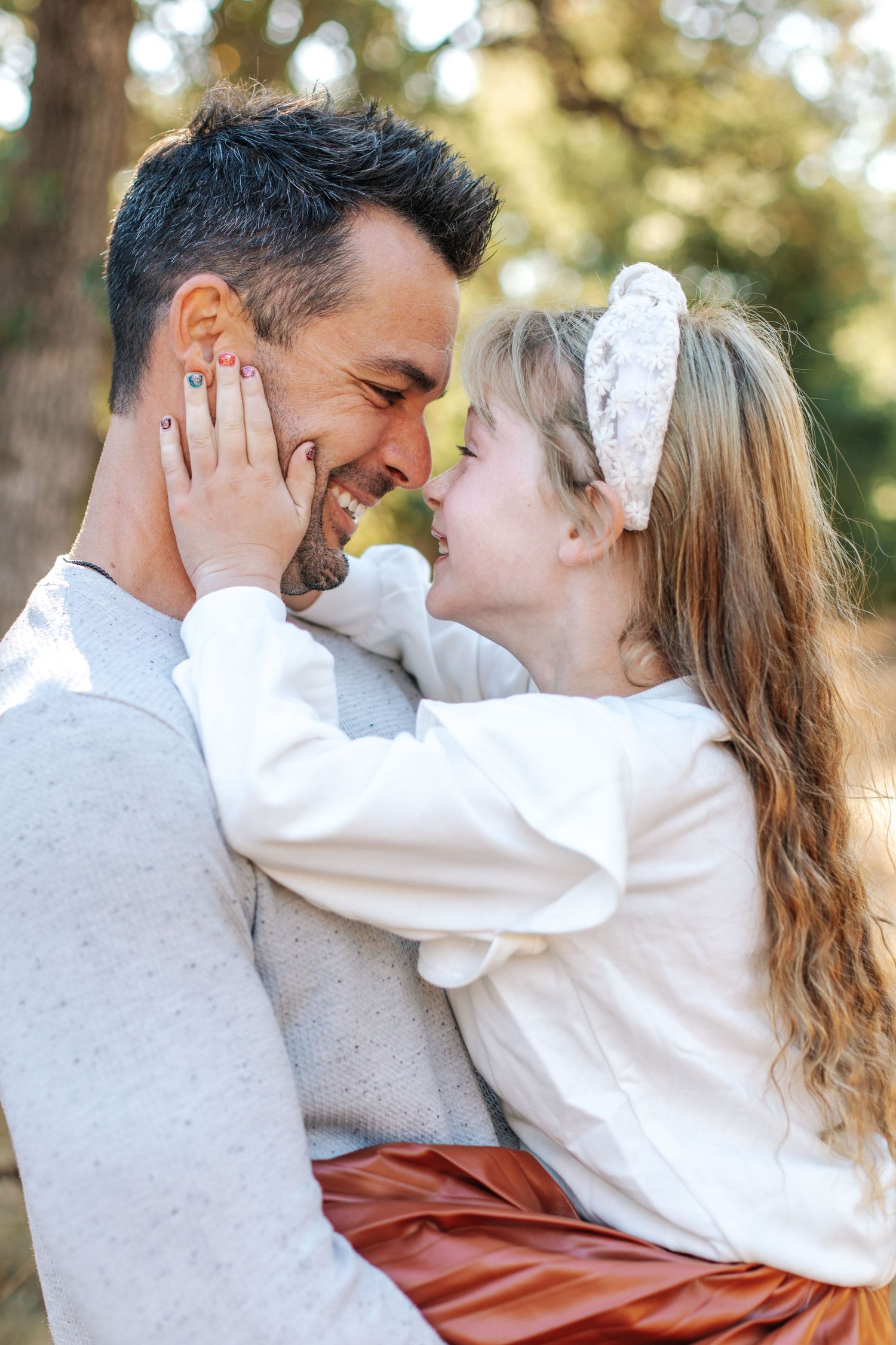 Smiling girl in white top tenderly holds fathers face in sunny outdoor setting.