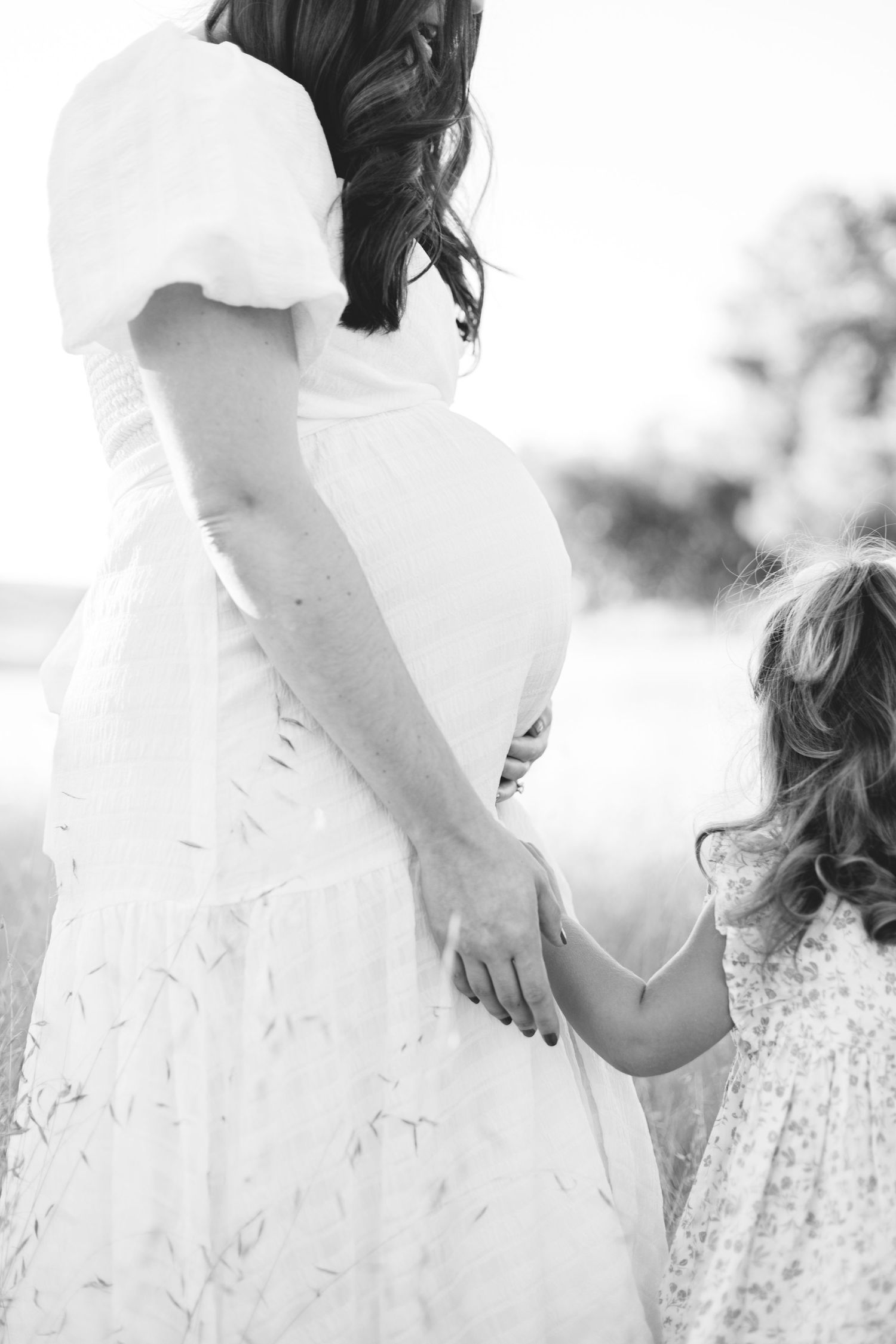 Black and white photo of toddler girl holding pregnant mothers hand outdoors.