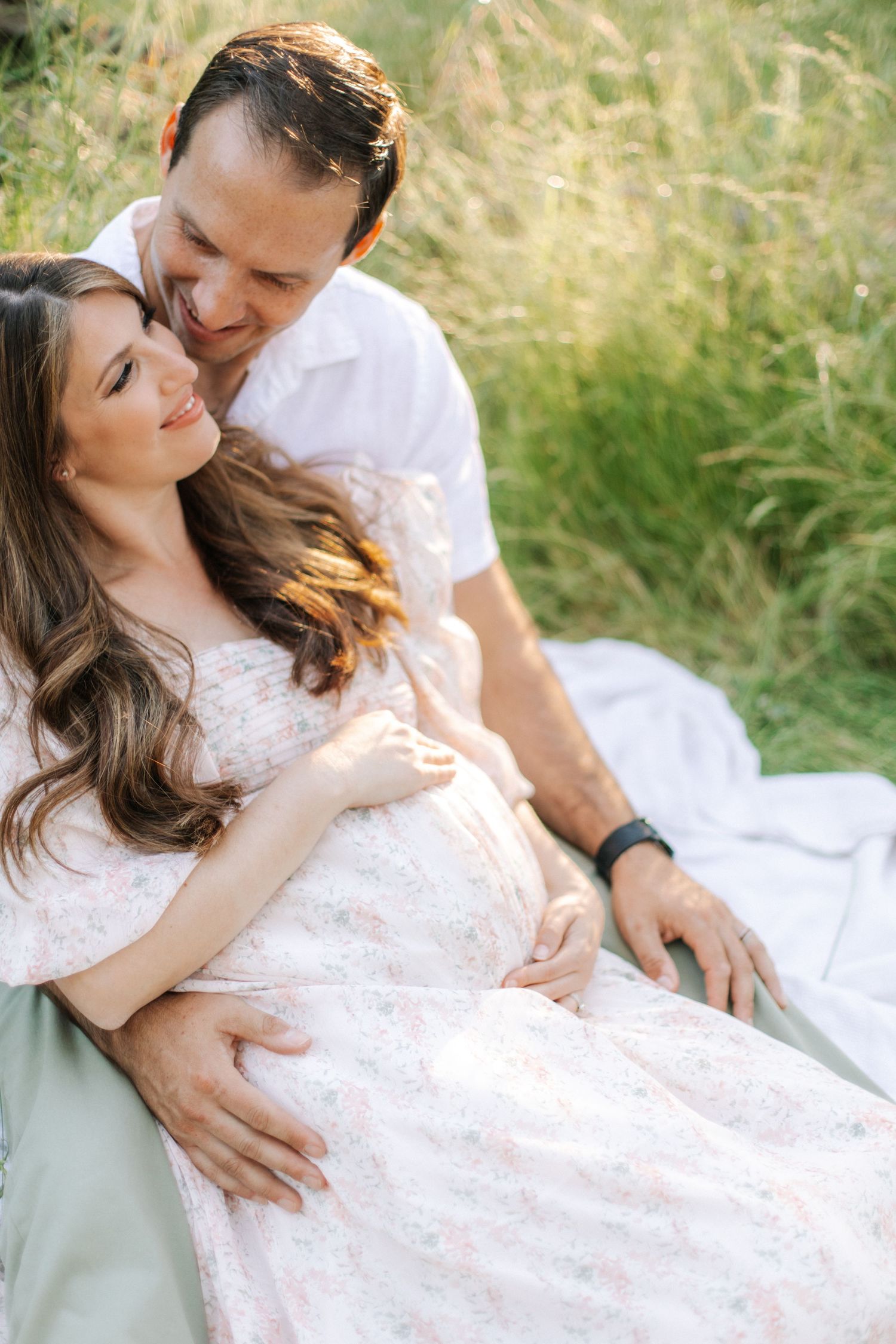 Couple sitting in grass field, man embracing pregnant woman in floral dress during maternity session.