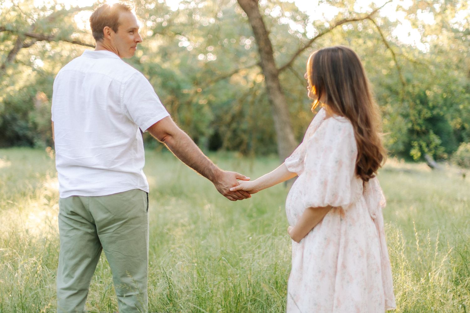 Pregnant couple holding hands walking through lush green meadow during golden hour maternity shoot.