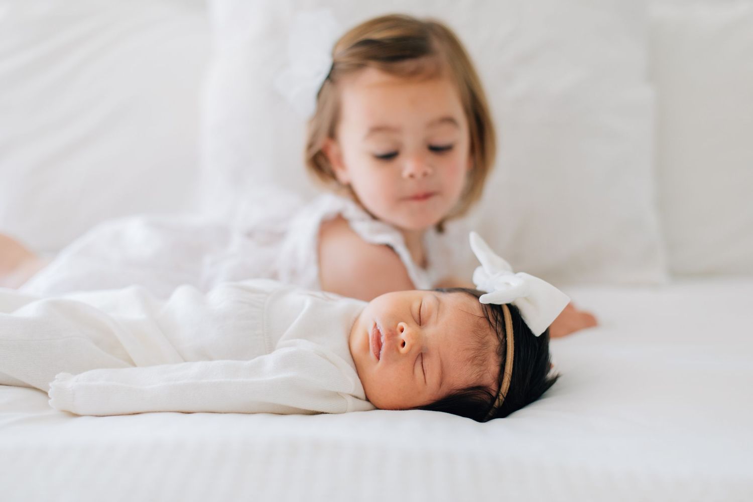 Peaceful swaddled newborn with bow headband sleeps on white bed as older toddler sister watches.