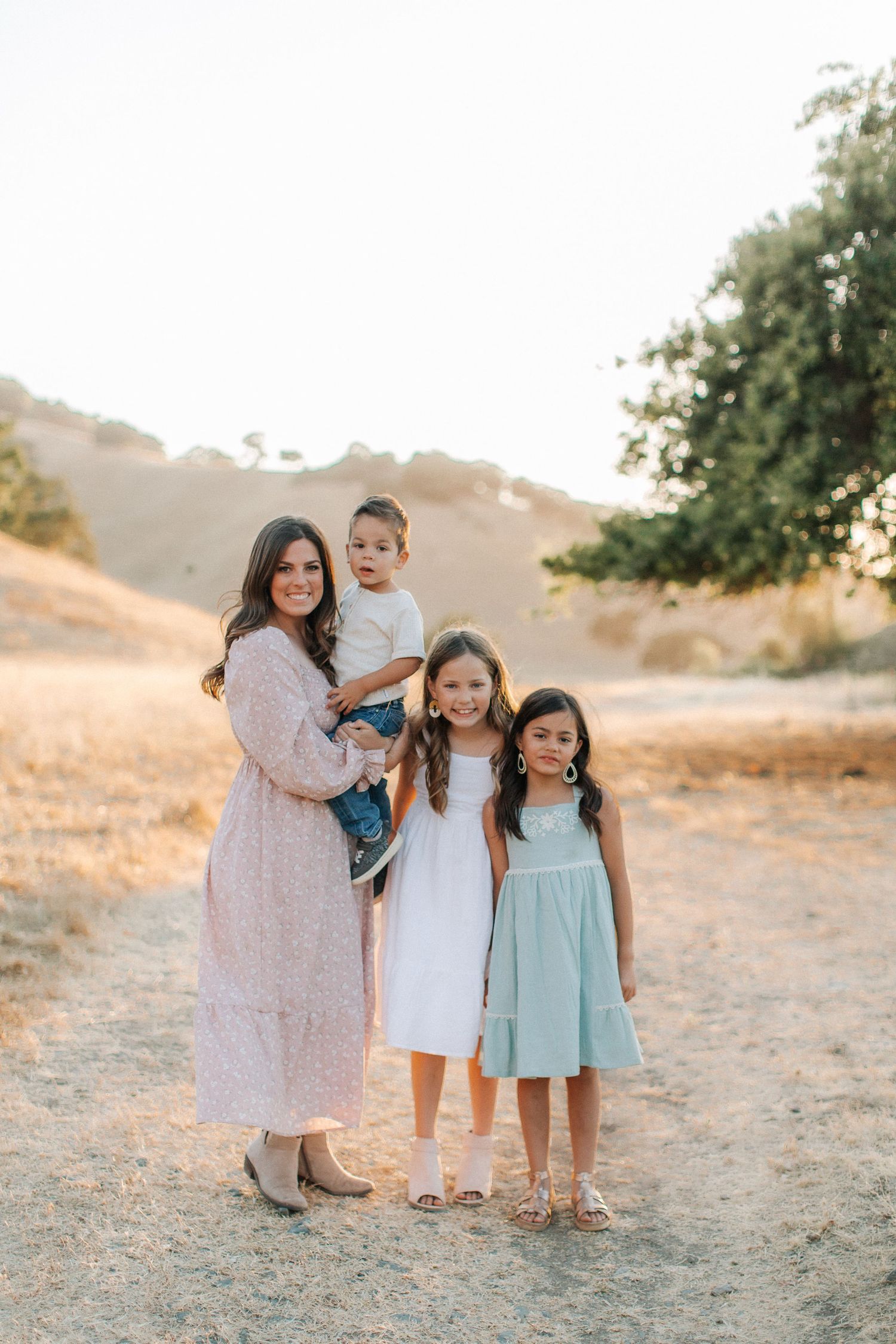 A mother in a pink dress holds her toddler son while posing with her two daughters outdoors on a sunny trail.