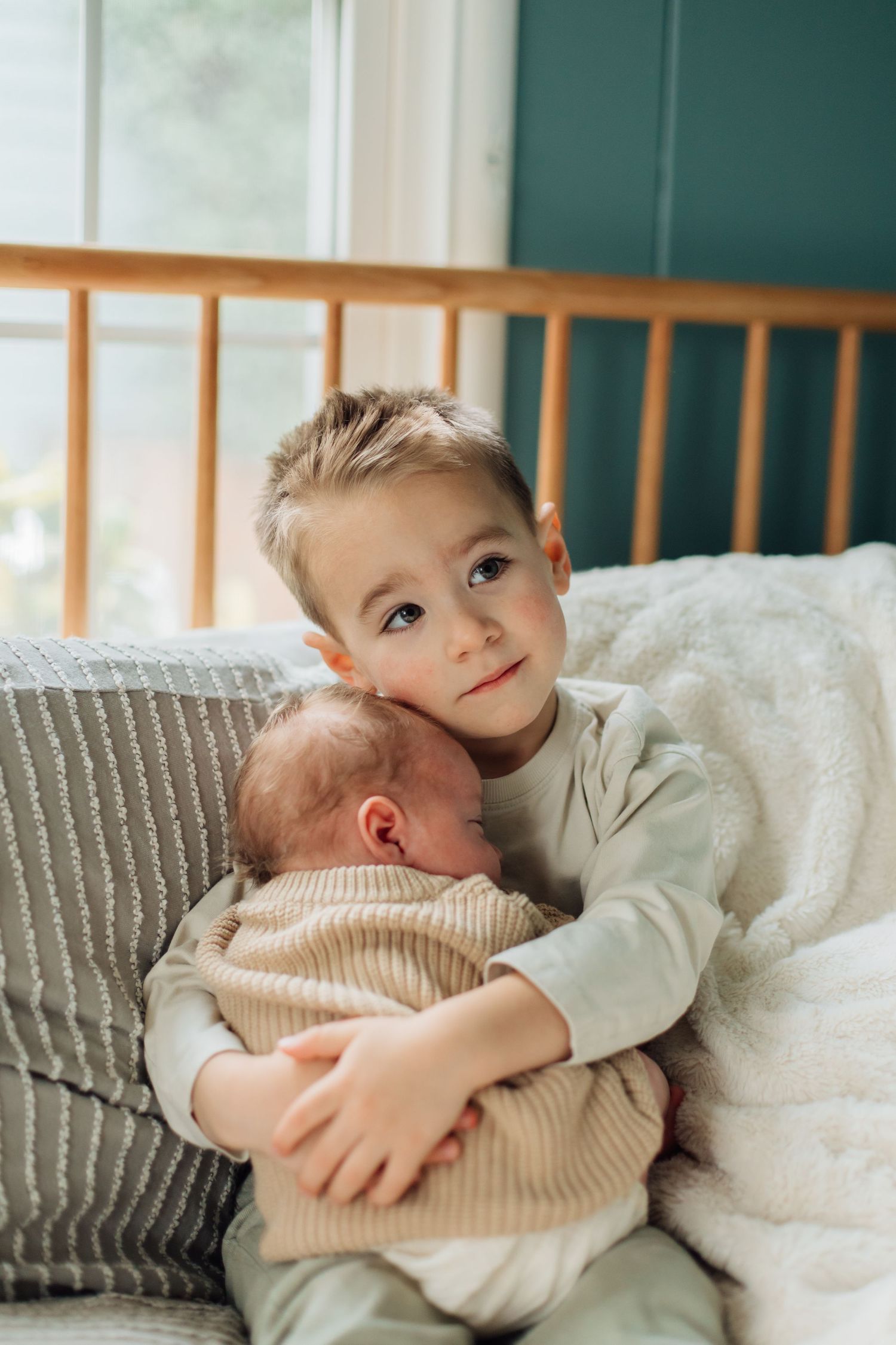 Young boy proudly holding newborn sibling while sitting on bed with teal wall background.