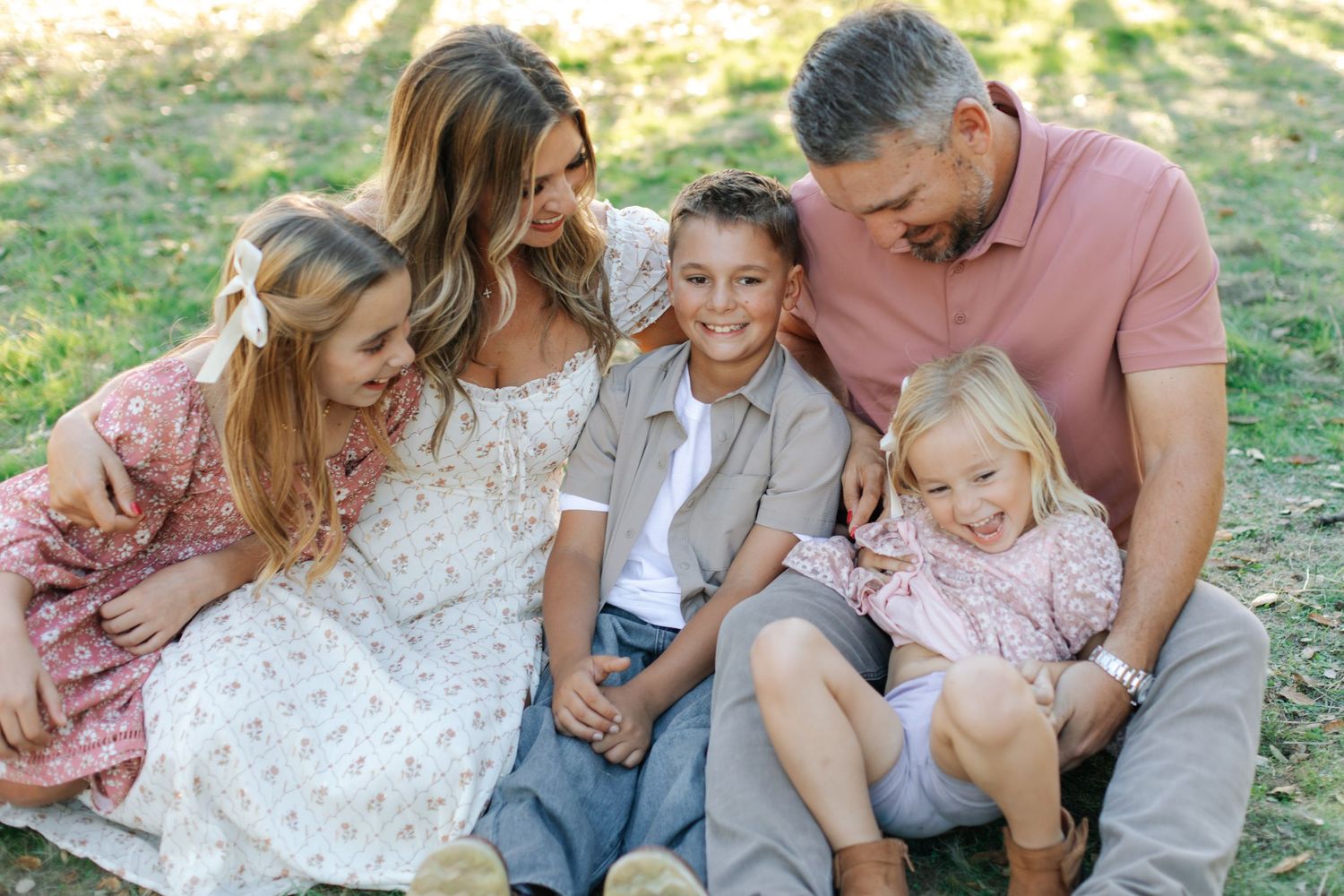 Happy family of five sitting together on grass in a sunny park, smiling and laughing.