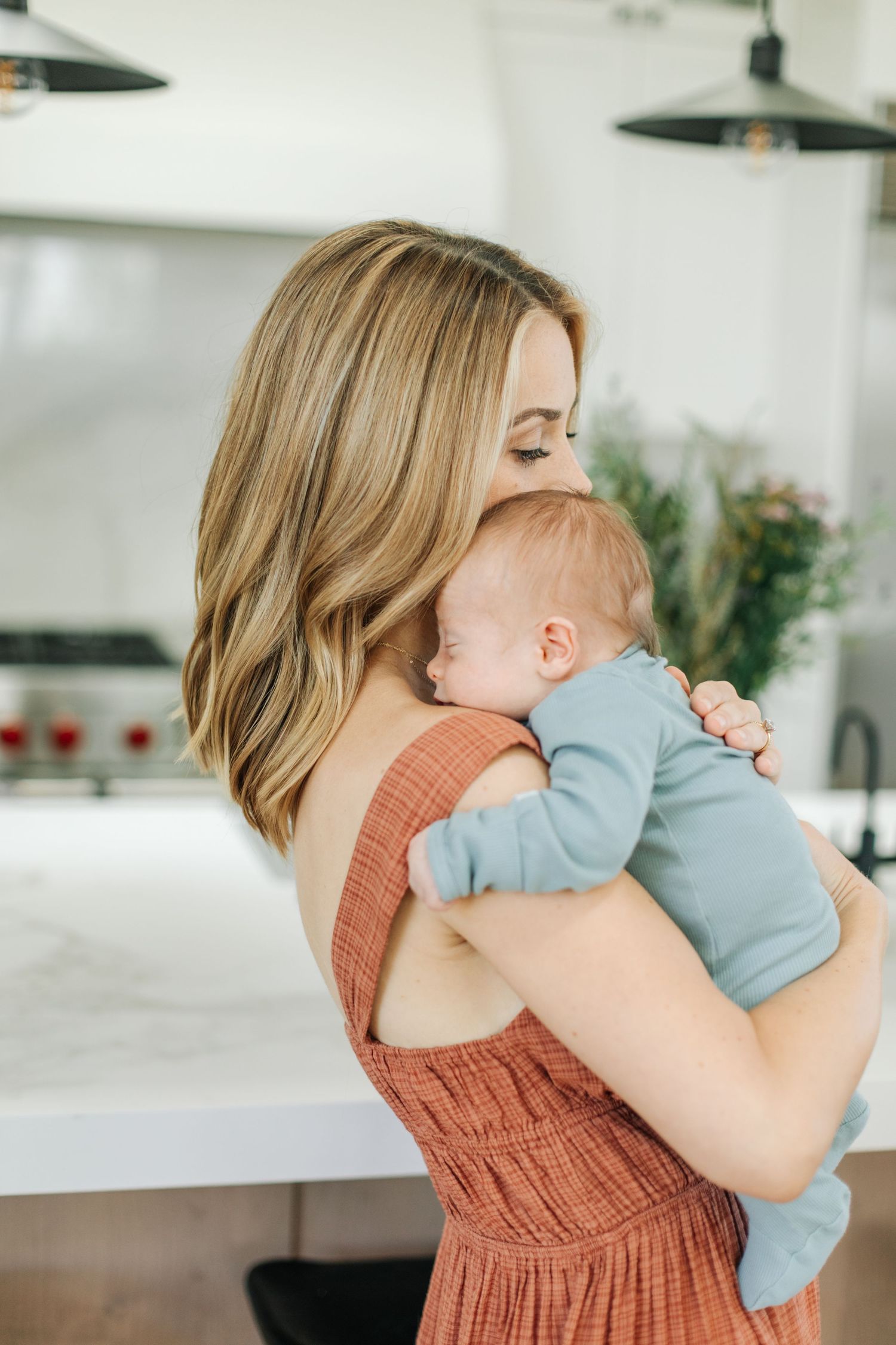 Mother in rust dress gently kissing her newborn baby on the head in modern kitchen.