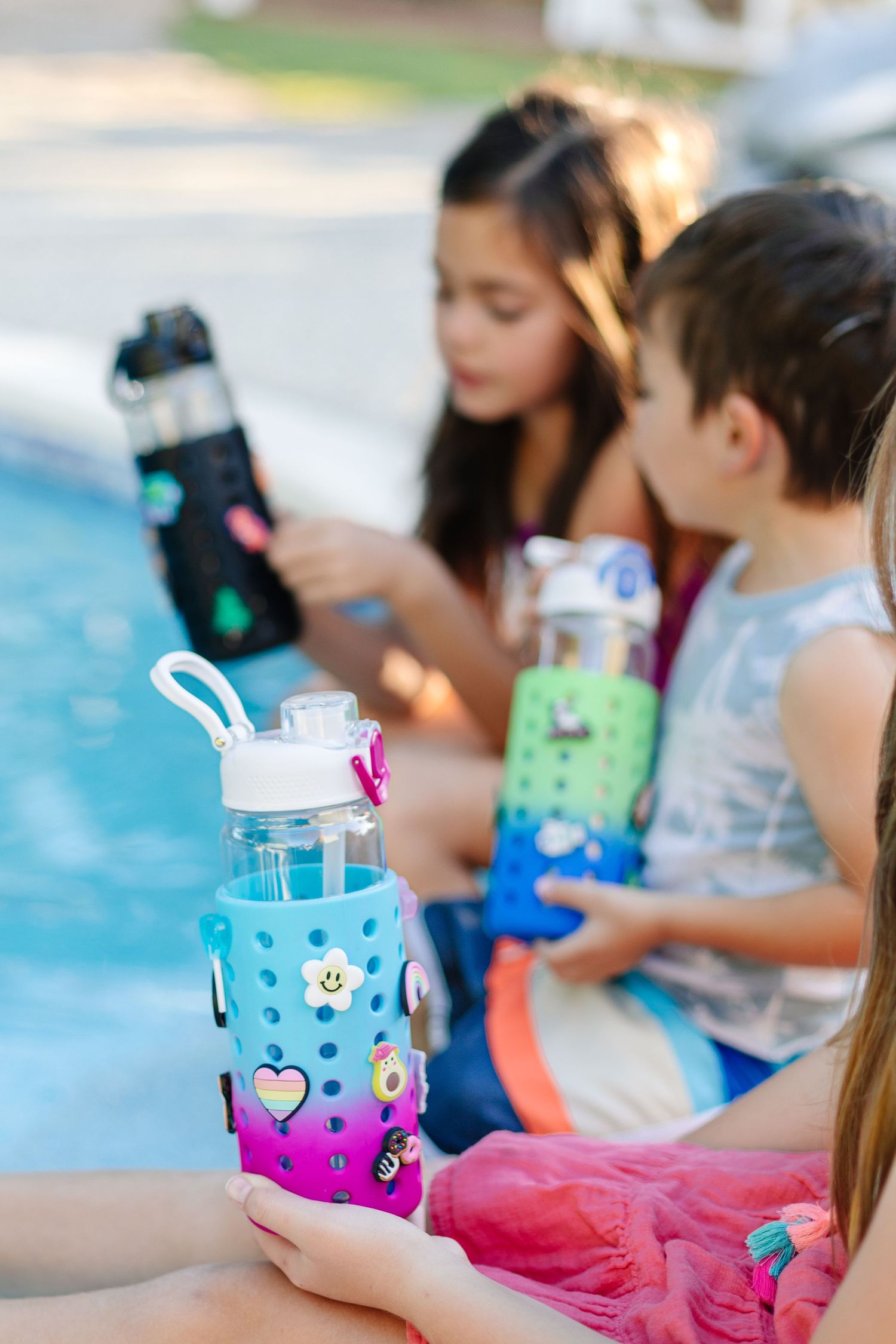 Three children holding colorful charm water bottles with silicone sleeves while sitting by a pool.