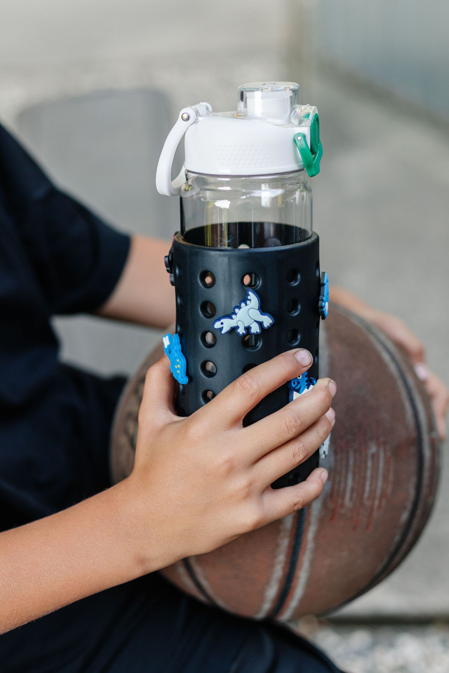 Child holding a black silicone-sleeved glass water bottle with dinosaur stickers near a basketball.