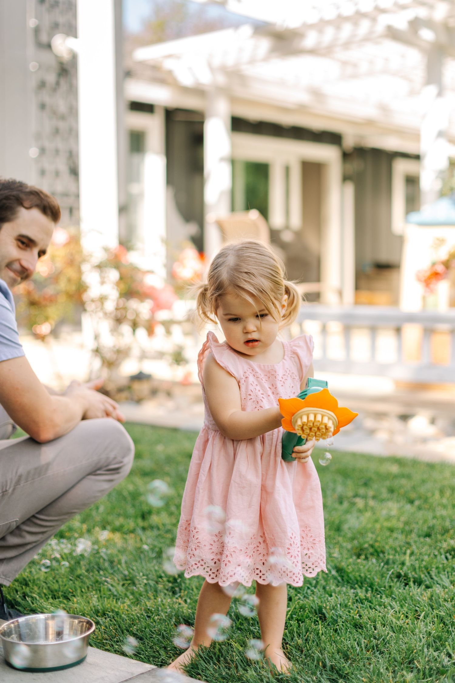 A toddler girl in a pink dress plays with a bubble gun in a backyard.