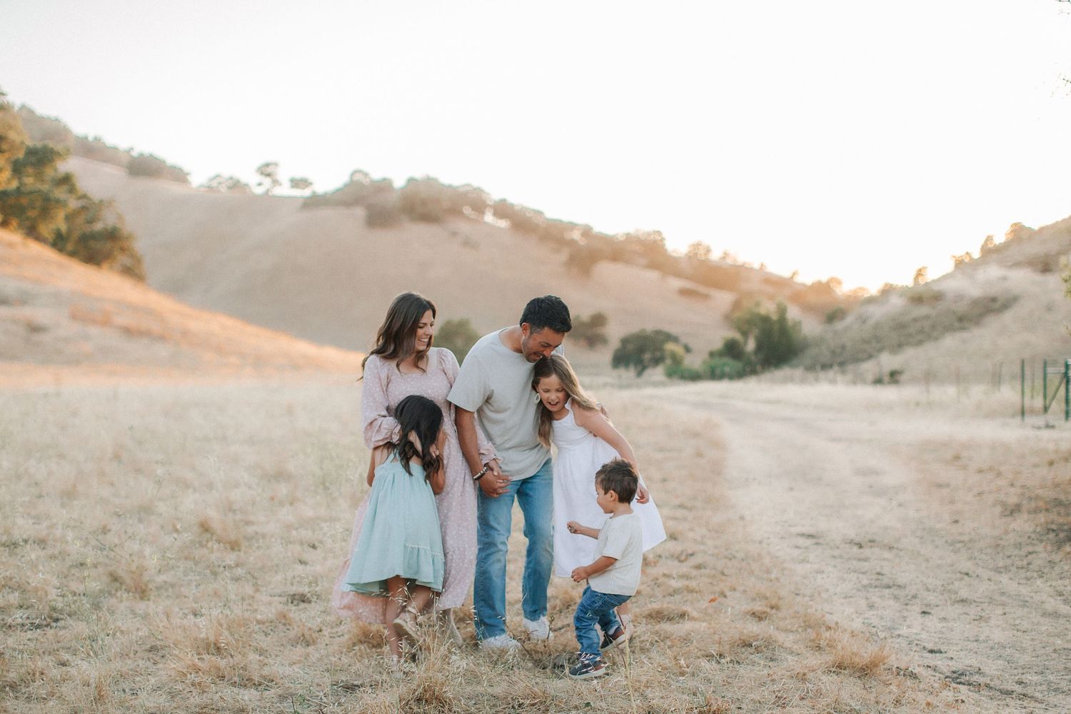 Joyful family of five gathered together on a sunlit dry grass path with rolling golden hills behind them.