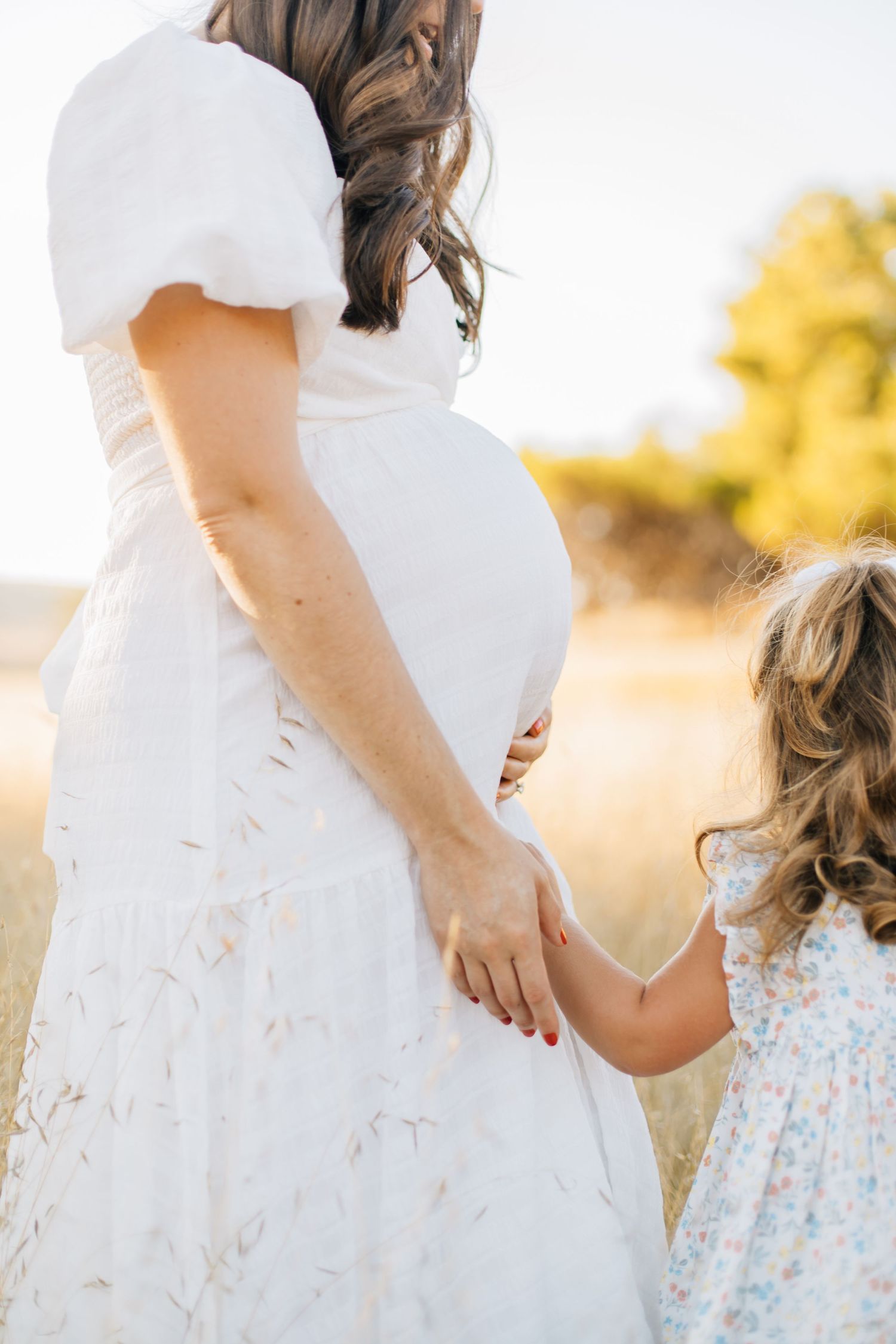 Pregnant woman in white dress holding hands with young daughter in sunny field.
