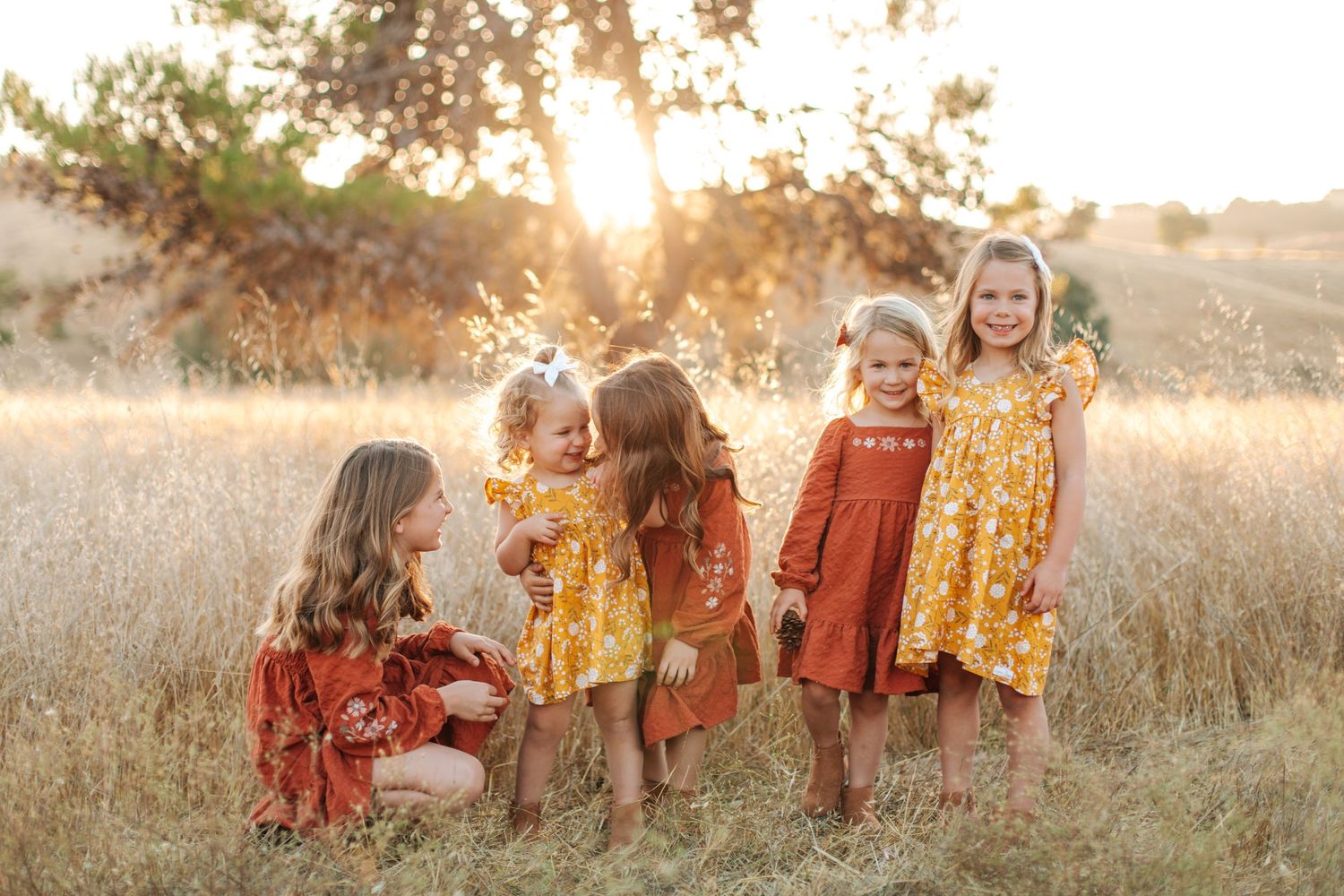 Five young girls in colorful floral dresses playing together in a golden sunlit field.