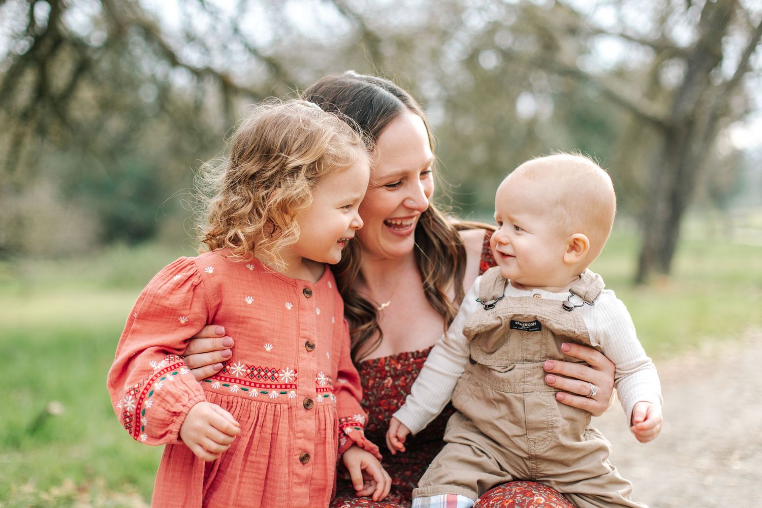 Happy mother smiling with curly-haired toddler girl and baby boy outdoors in a park.