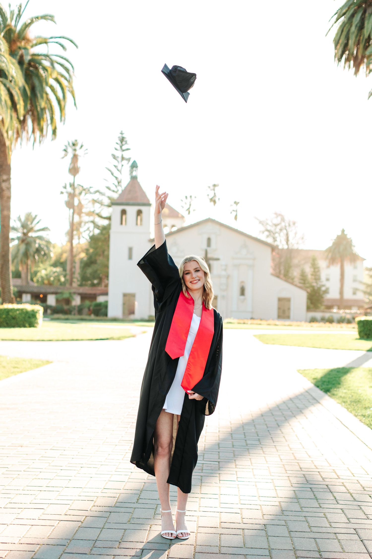 Joyful blonde graduate in black gown tossing cap in air on brick path near white mission church.