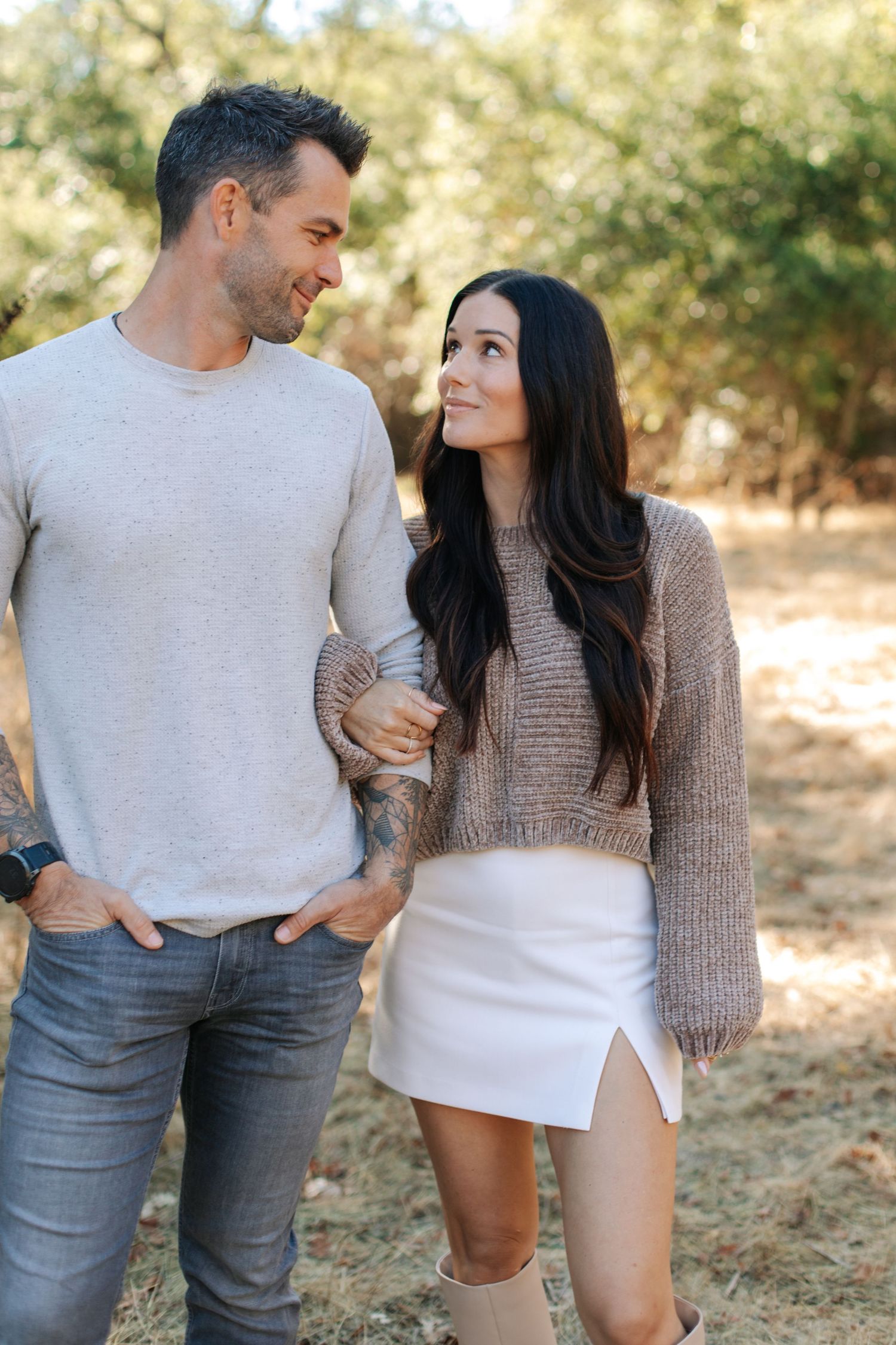 Stylish couple walking arm in arm through a sunny tree-lined outdoor path.