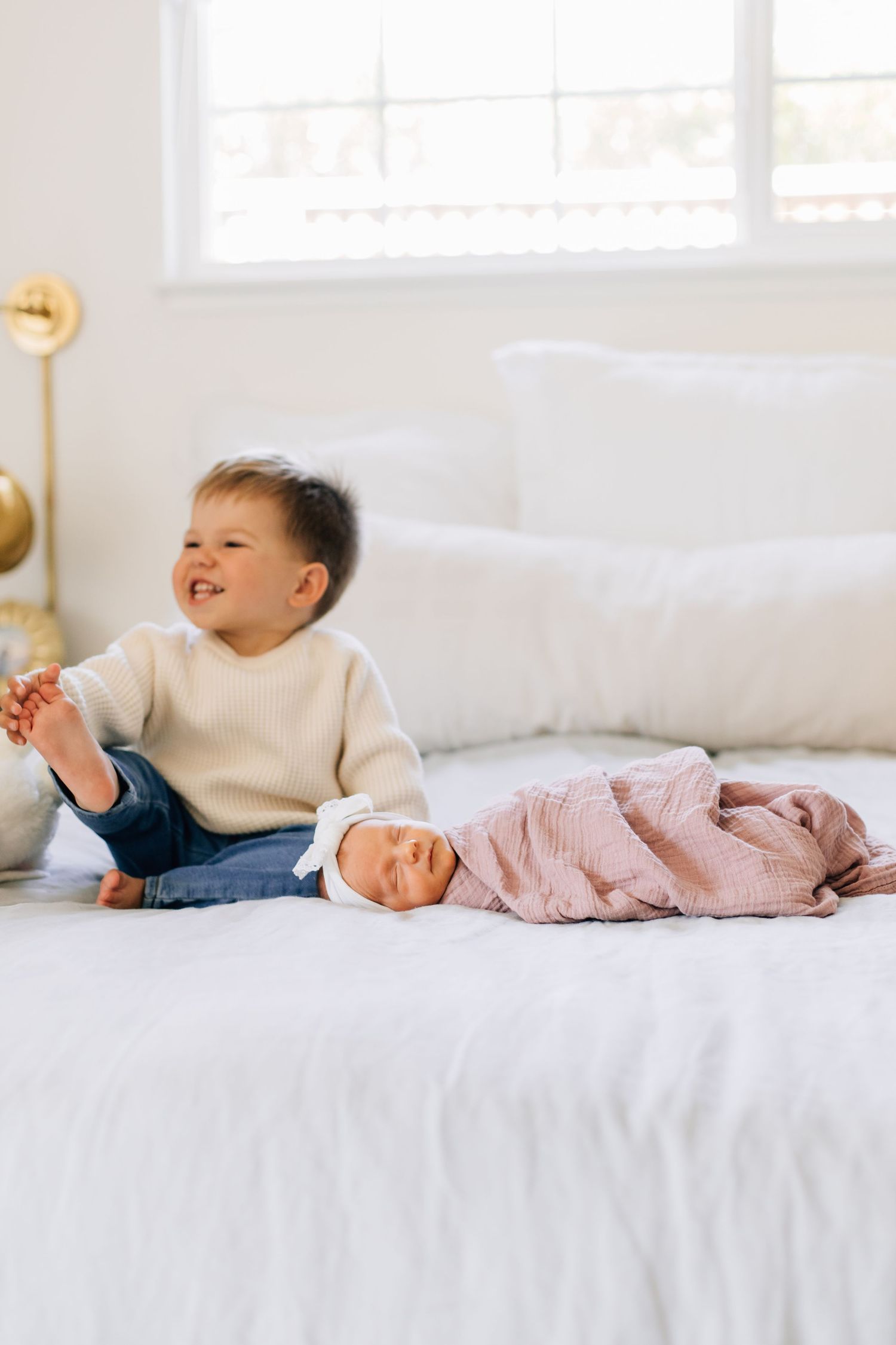 Smiling toddler boy sits on white bed next to sleeping newborn sister wrapped in pink swaddle.