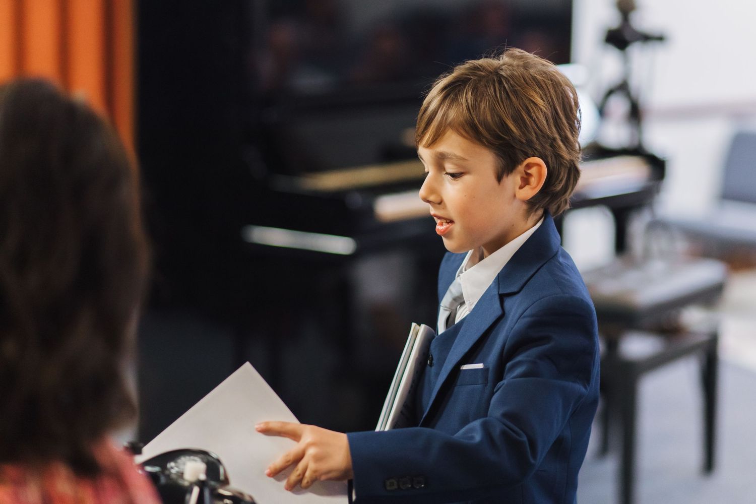 A boy in a navy suit reads sheet music at a piano recital.